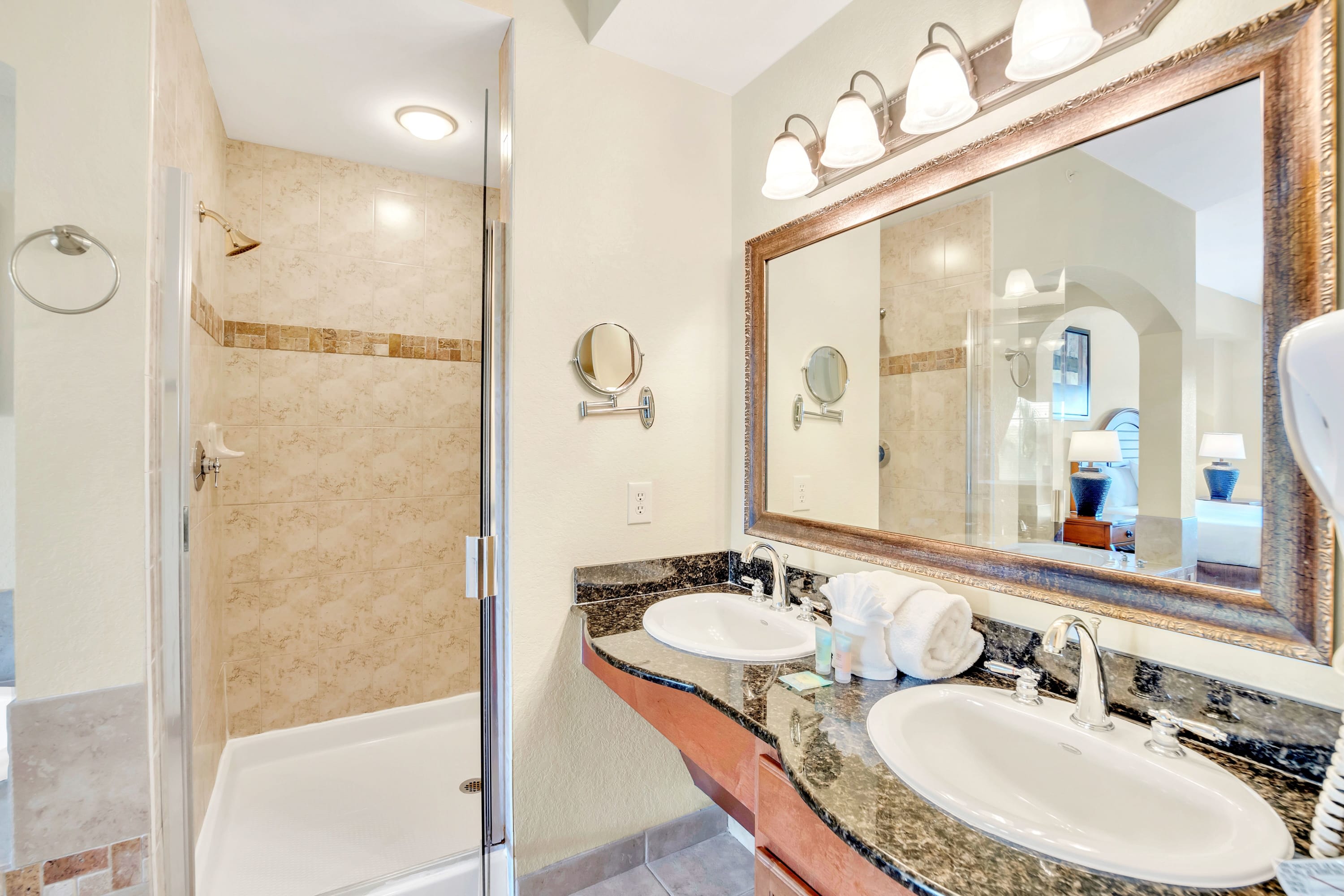 Master bathroom with a double vanity featuring granite countertops and dark wood cabinets, a large ornate mirror, and a walk-in shower with tiled walls and a glass door.
