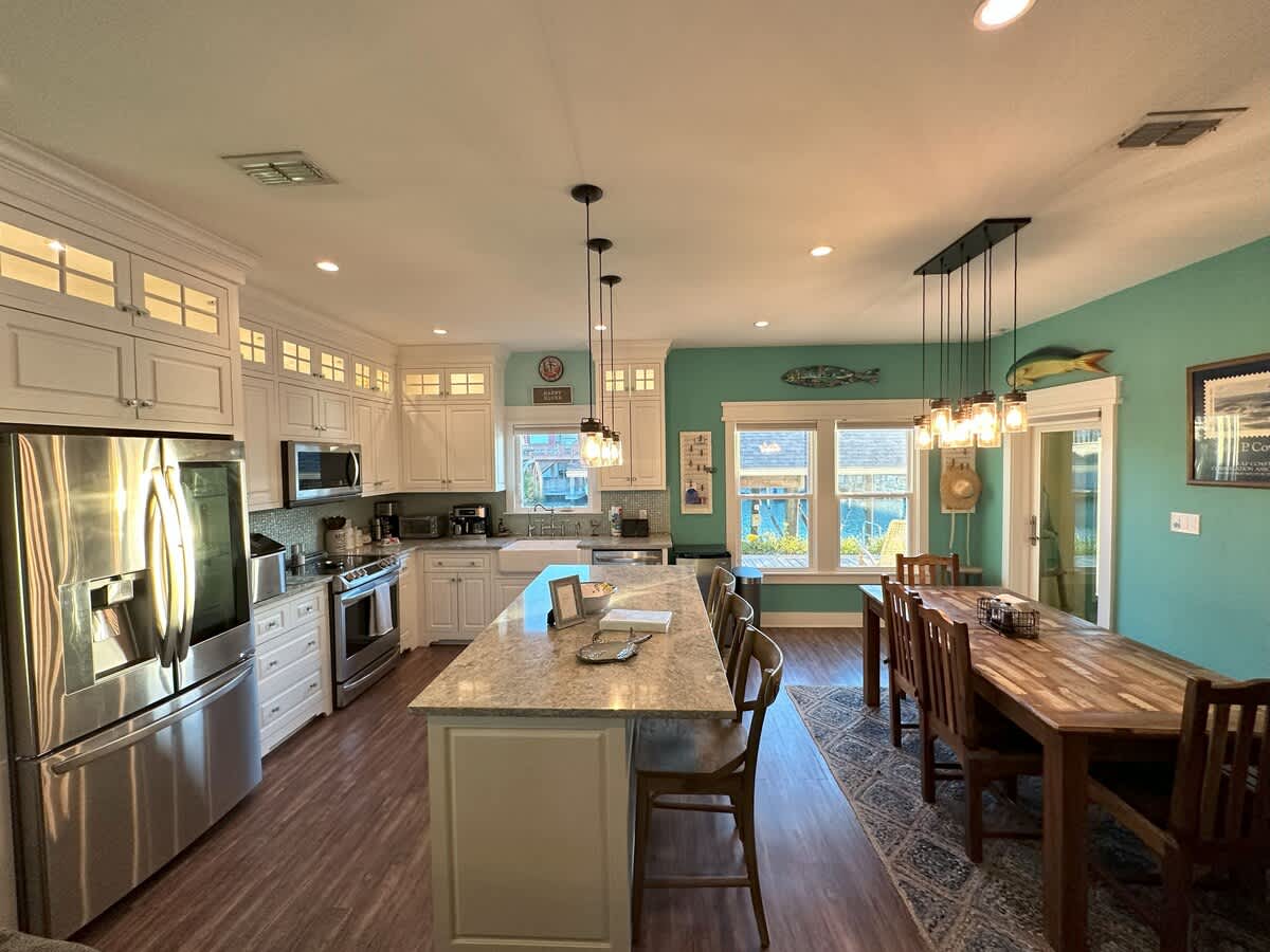 Modern kitchen featuring white shaker cabinets with glass inserts, stainless steel appliances, a large granite island with bar stools, and a farmhouse sink, illuminated by pendant and recessed lighting.
