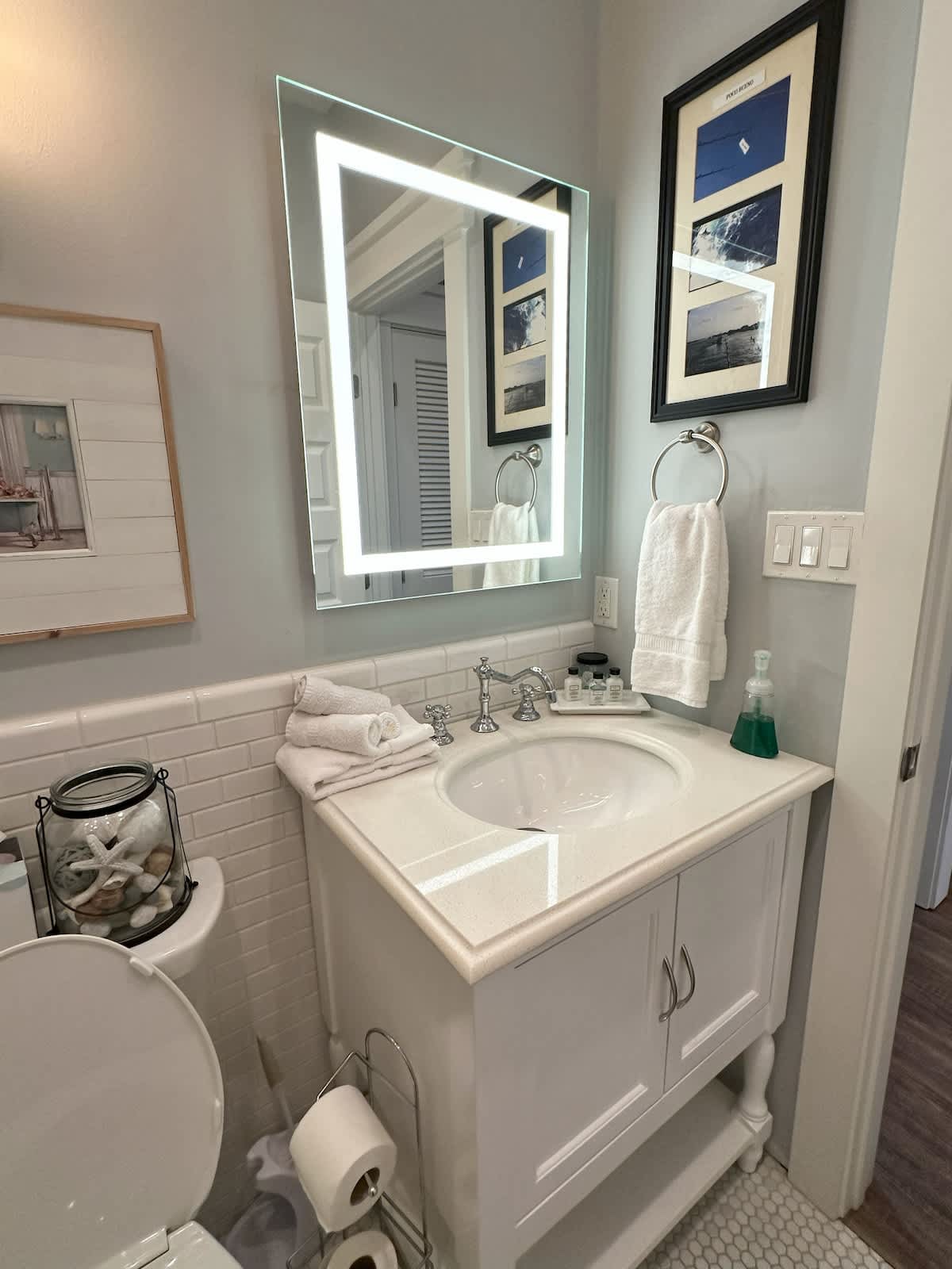 A bright bathroom features a white vanity with a rectangular undermount sink, a large illuminated mirror, and classic white subway tile wainscoting.