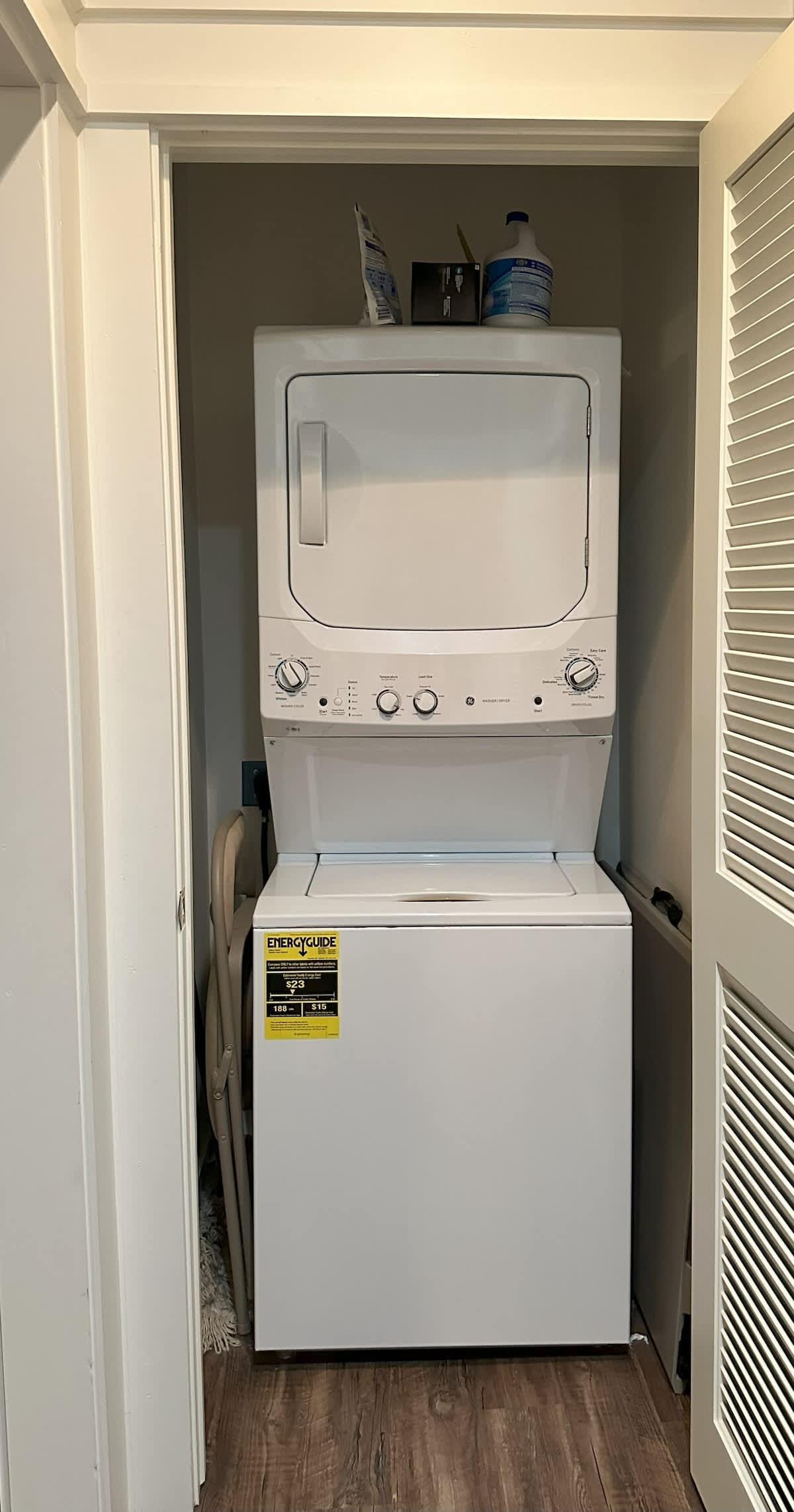 A stacked white washer and dryer unit is neatly tucked into a closet with louvered bifold doors, indicating a dedicated laundry area. Cleaning supplies are visible on a shelf above the unit.