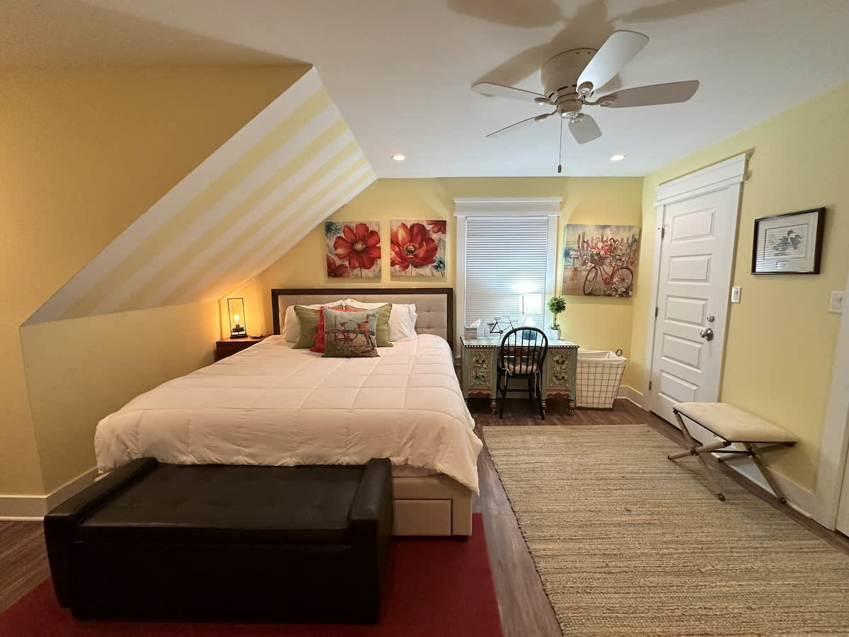 A bright bedroom with yellow walls and a sloped ceiling, featuring a queen-sized bed with a padded headboard, a decorative desk with a chair, and a window with blinds. Artworks with floral and bicycle themes adorn the walls.