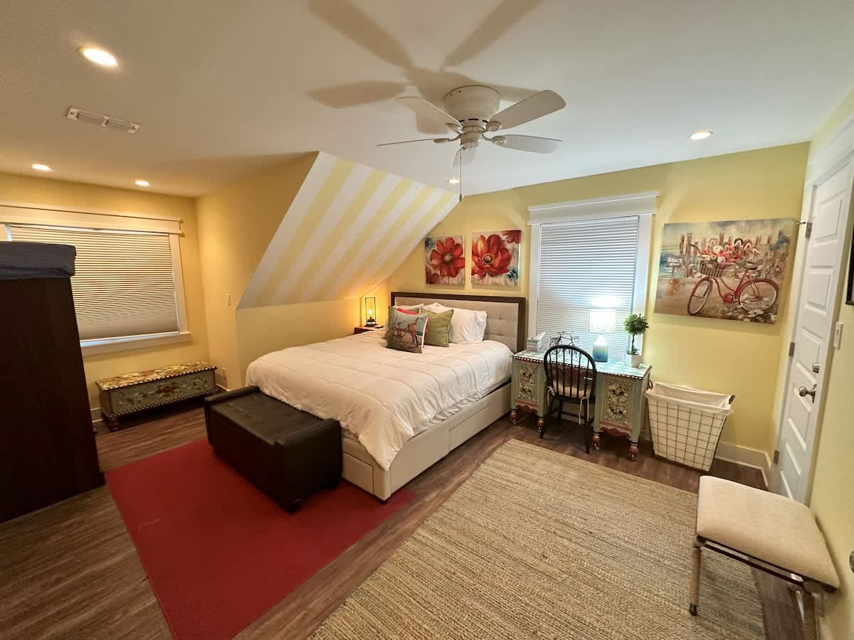 A spacious bedroom with yellow walls and a sloped ceiling, featuring a queen-sized bed, a desk with a chair, and a large area rug. Two windows with blinds and various artworks are visible, along with a dark storage ottoman.