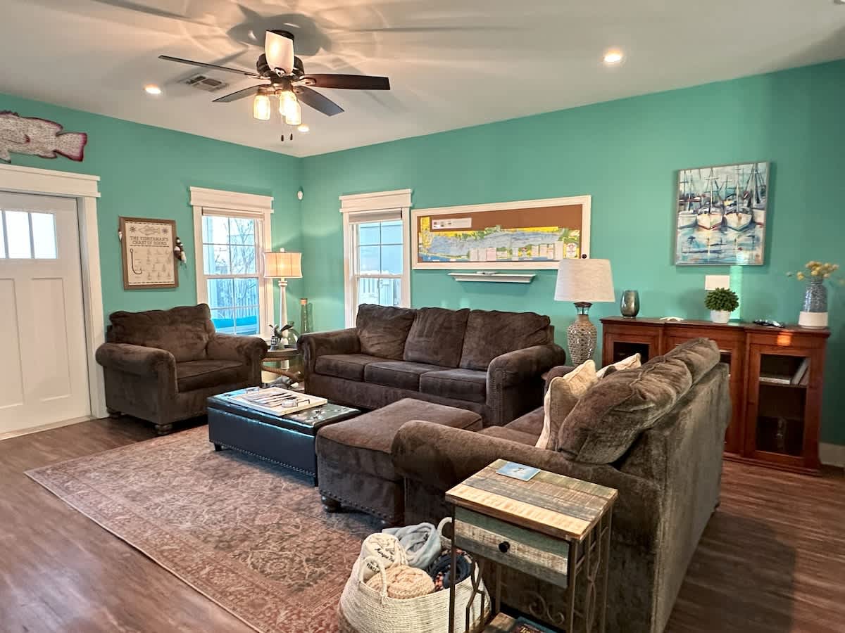 Spacious living room with vibrant teal walls, comfortable brown seating including two sofas and an armchair, an ottoman coffee table, and multiple windows, creating a bright and welcoming atmosphere.