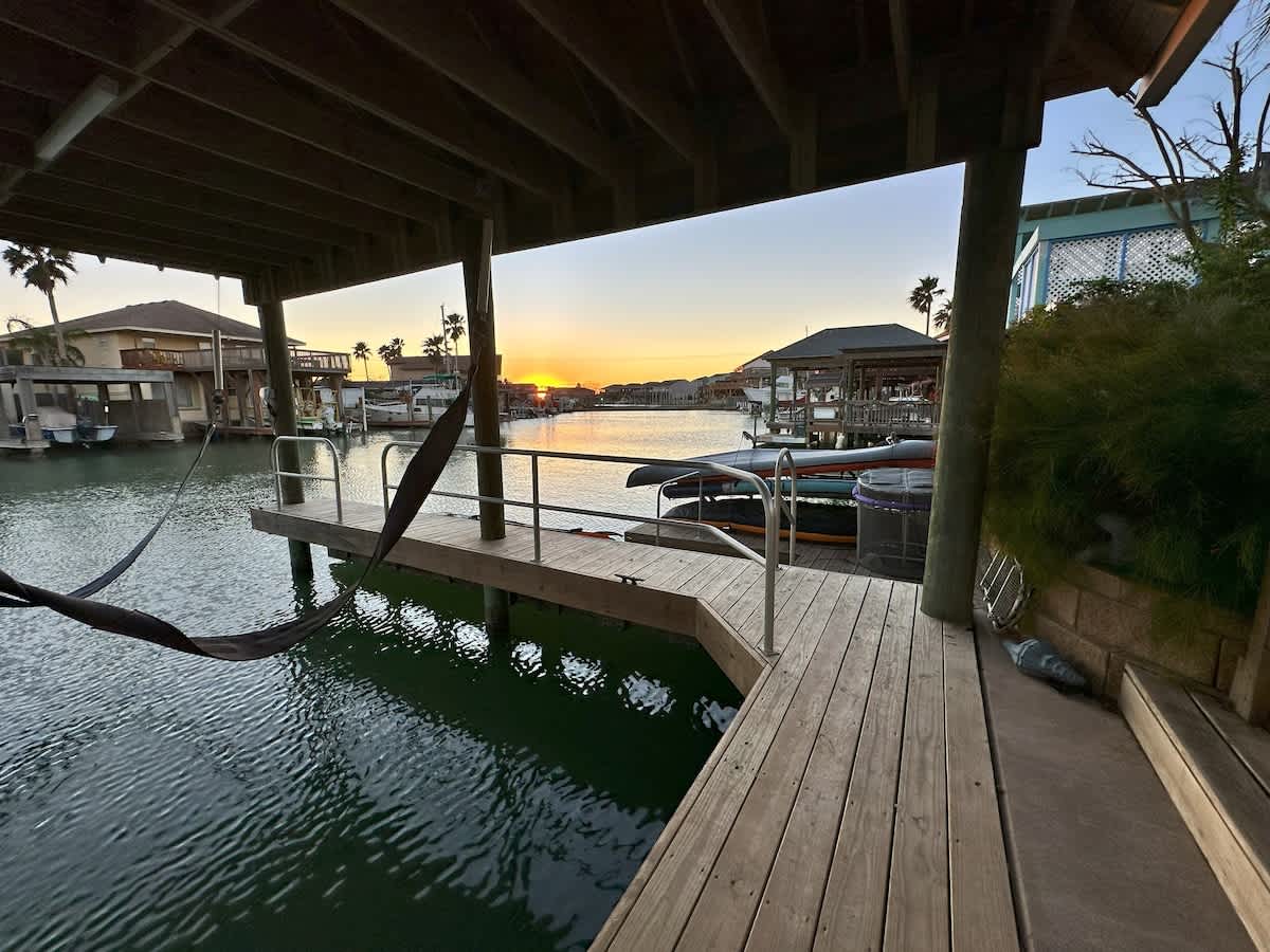 A wooden boat dock extends into the serene canal, featuring a hanging hammock and offering a beautiful view of the sunset painting the sky over the water in the distance.
