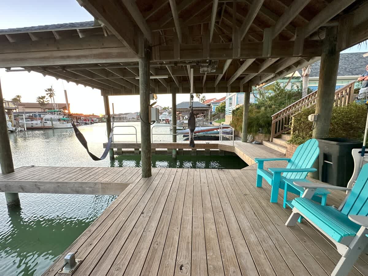 Under a covered wooden boat dock, a comfortable hammock and two light blue Adirondack chairs invite relaxation, overlooking the calm canal water with other houses in the background.