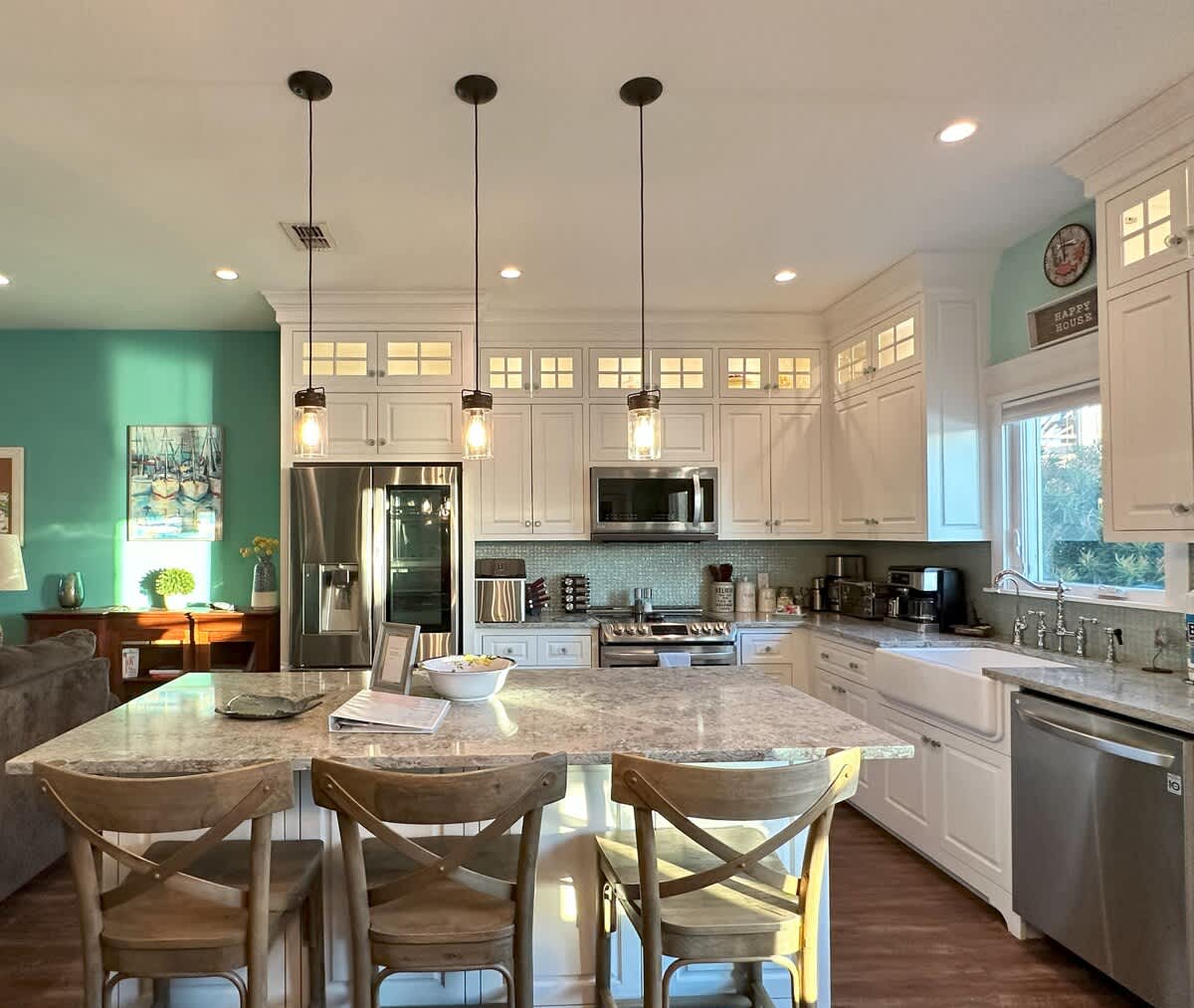 Bright kitchen with white shaker cabinets, stainless steel appliances, a large granite island with three wooden bar stools, and a view towards the dining area and living room, all with hardwood flooring.