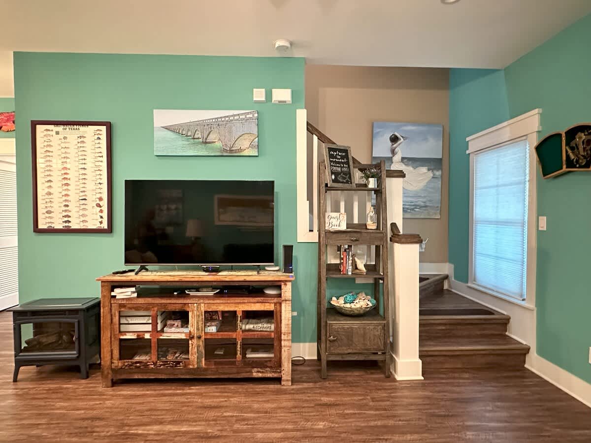 View of the living room showing the large flat-screen TV on a rustic wooden console, teal walls, and a staircase leading to another level, with nautical-themed artwork and decor.