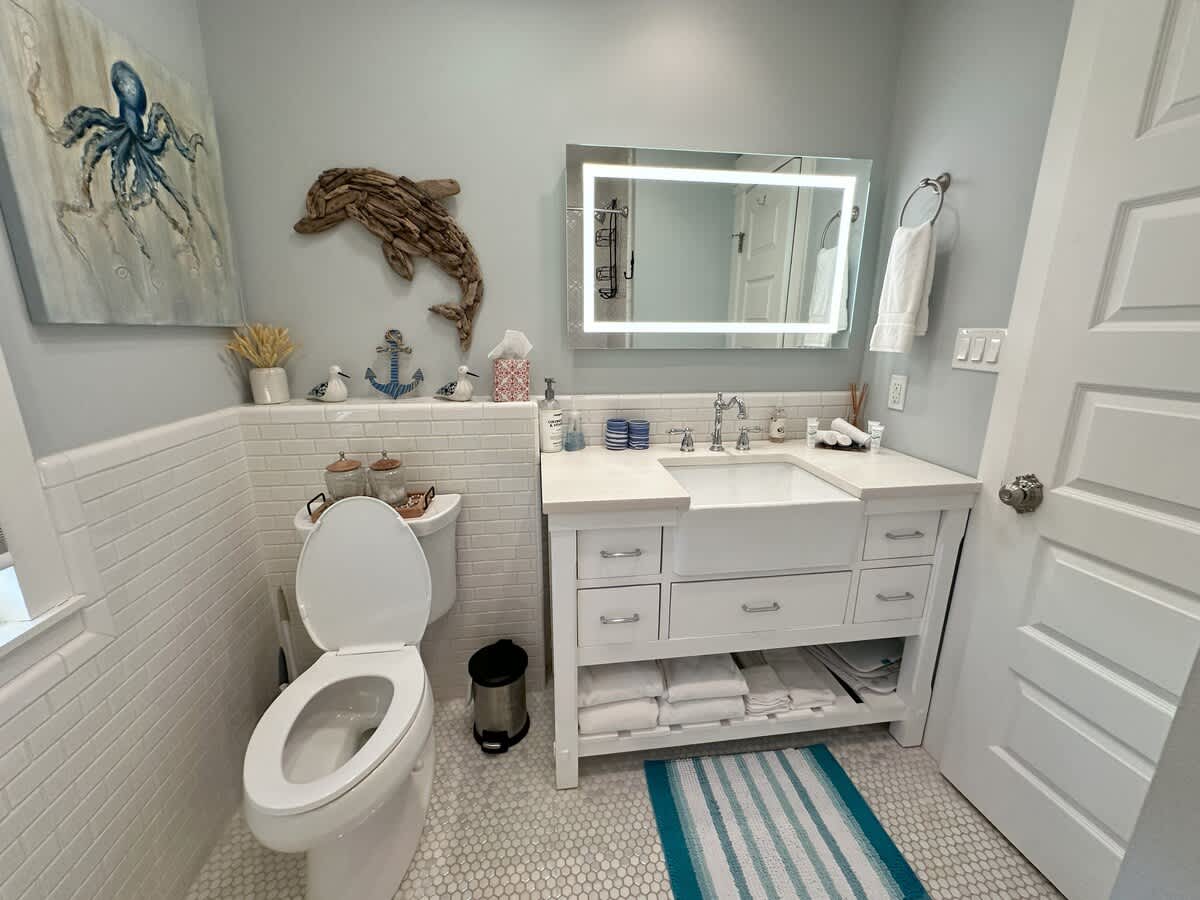 A detailed view of a bathroom with light blue walls, showcasing a white vanity with a farmhouse sink, a toilet, and nautical-themed decor including an octopus painting and a wooden dolphin. White hexagonal mosaic tiles cover the floor.