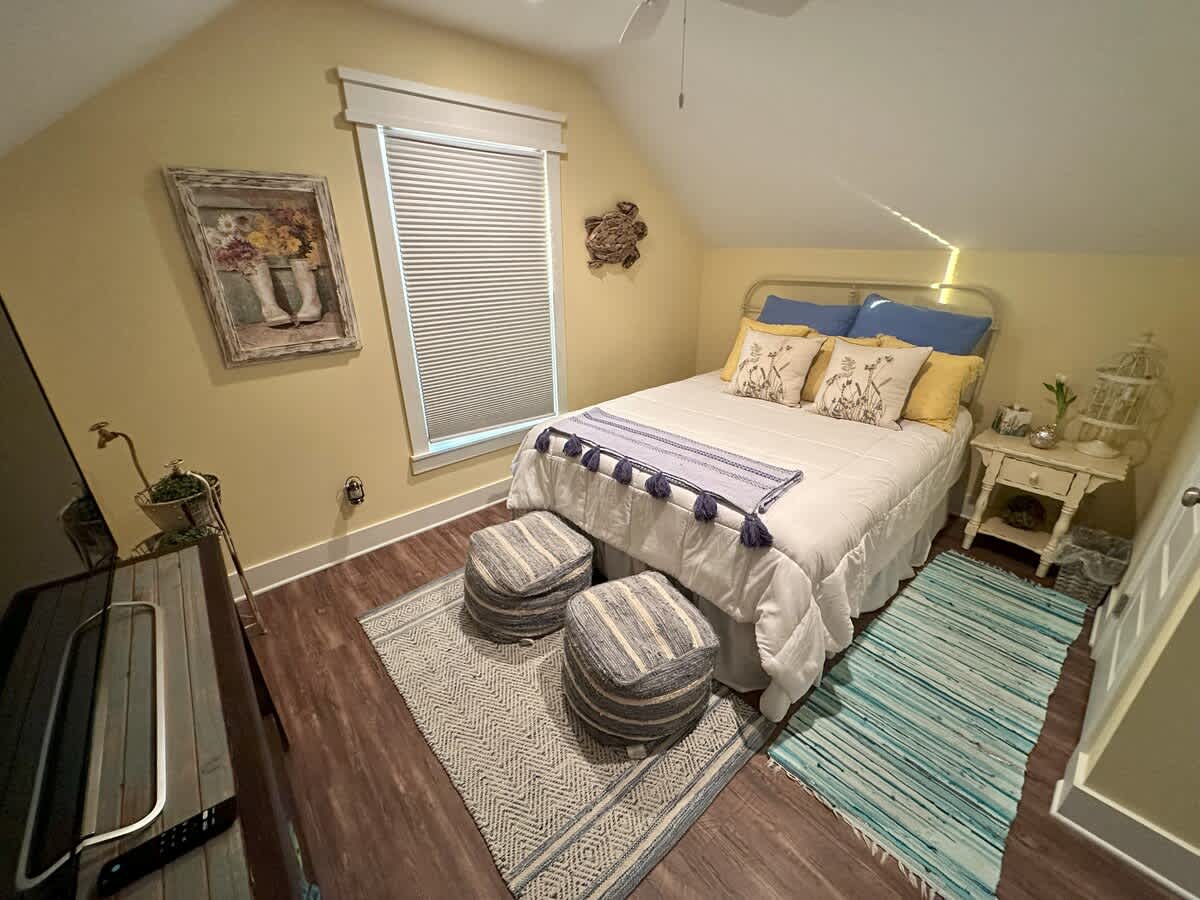 A cozy bedroom with light yellow walls and a sloped ceiling, featuring a white metal-frame bed with blue and yellow pillows, two striped ottomans, and a patterned area rug on dark wood-look flooring. A window with blinds is visible.