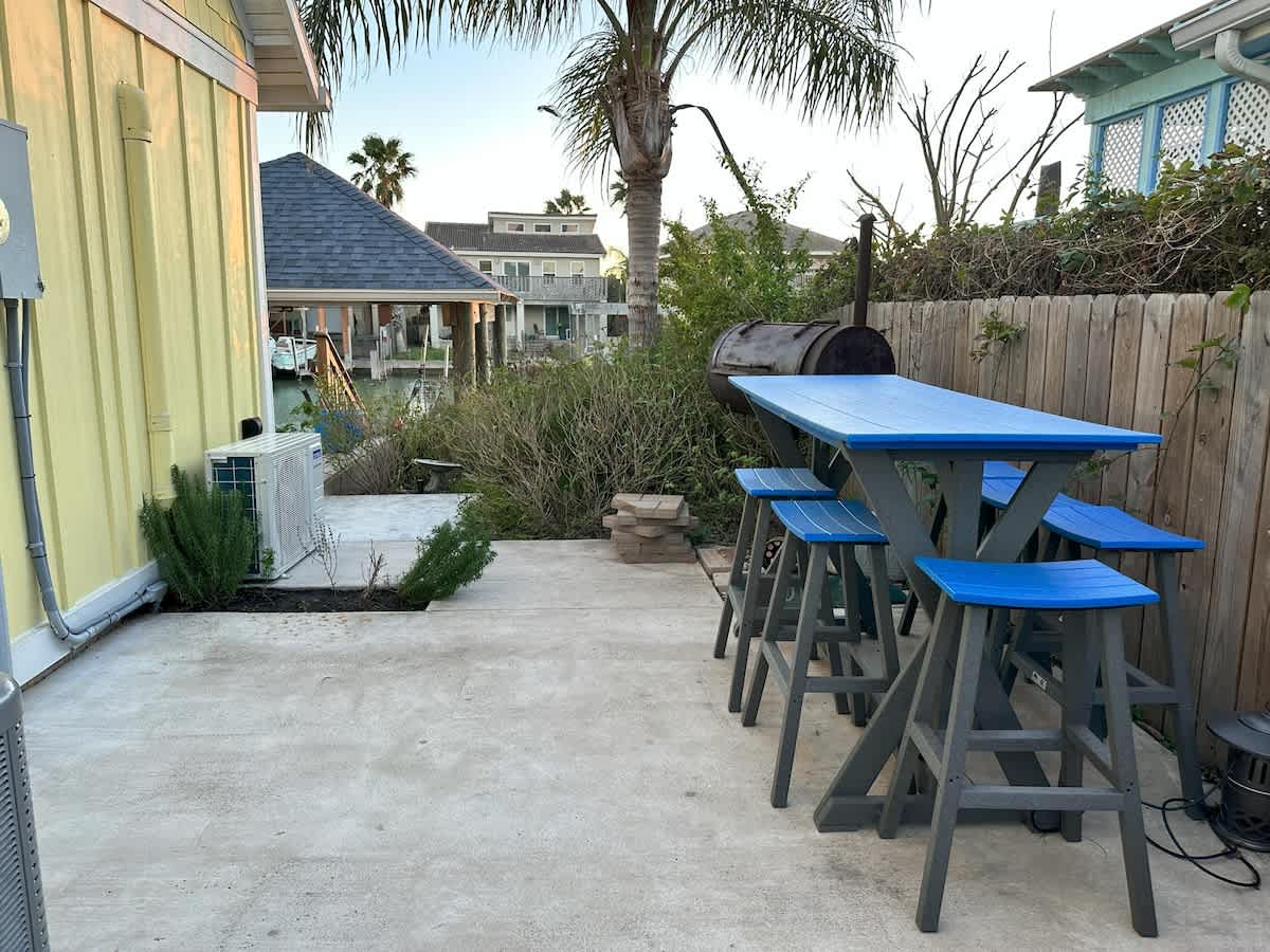 A concrete patio area features a blue picnic table with matching stools and a large BBQ grill, bordered by a wooden fence and lush green landscaping.