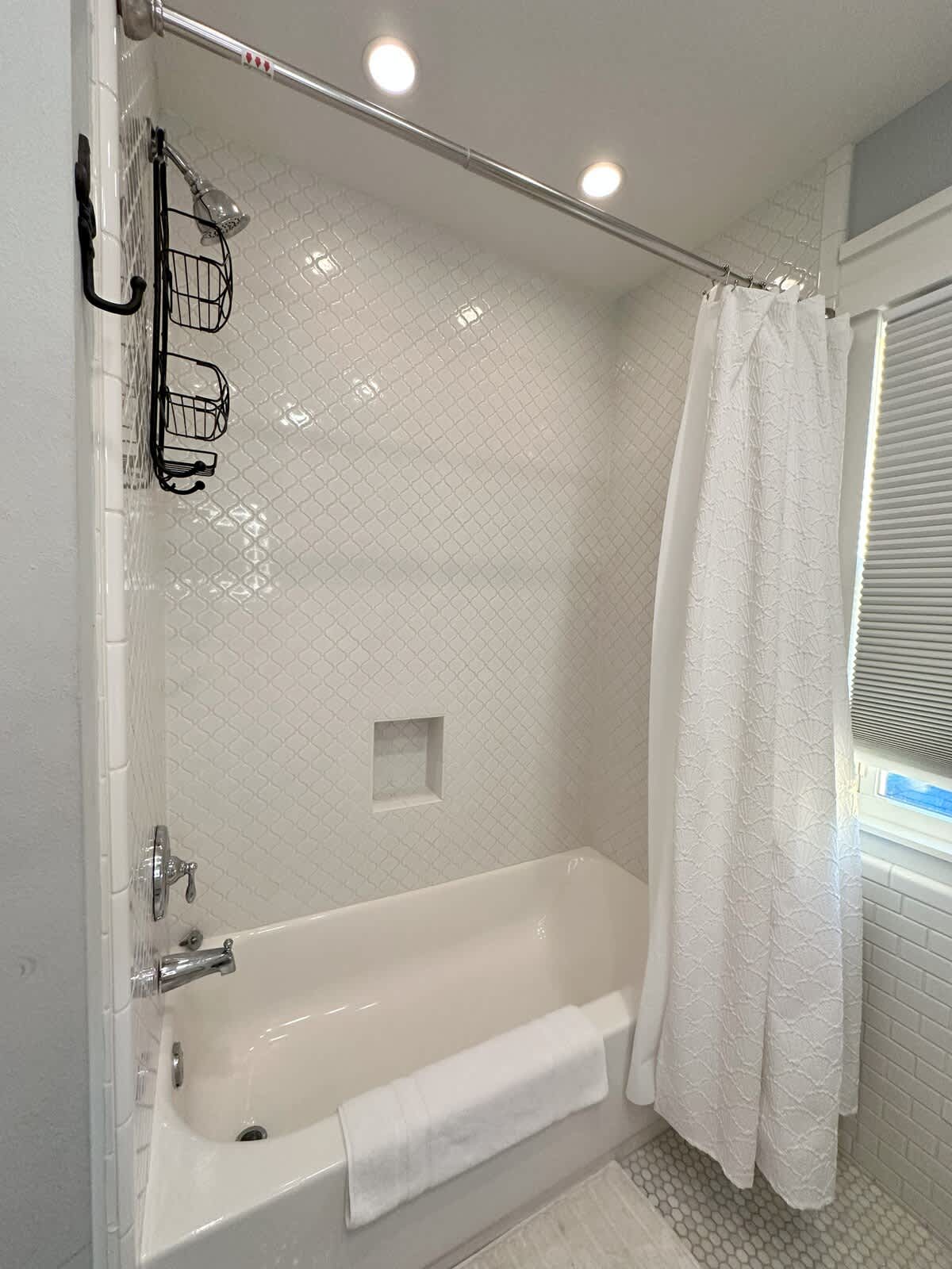 A white bathtub with a shower curtain in a bathroom featuring white diamond-patterned wall tiles and recessed lighting. A shower caddy hangs on the wall, and white hexagonal mosaic tiles cover the floor.