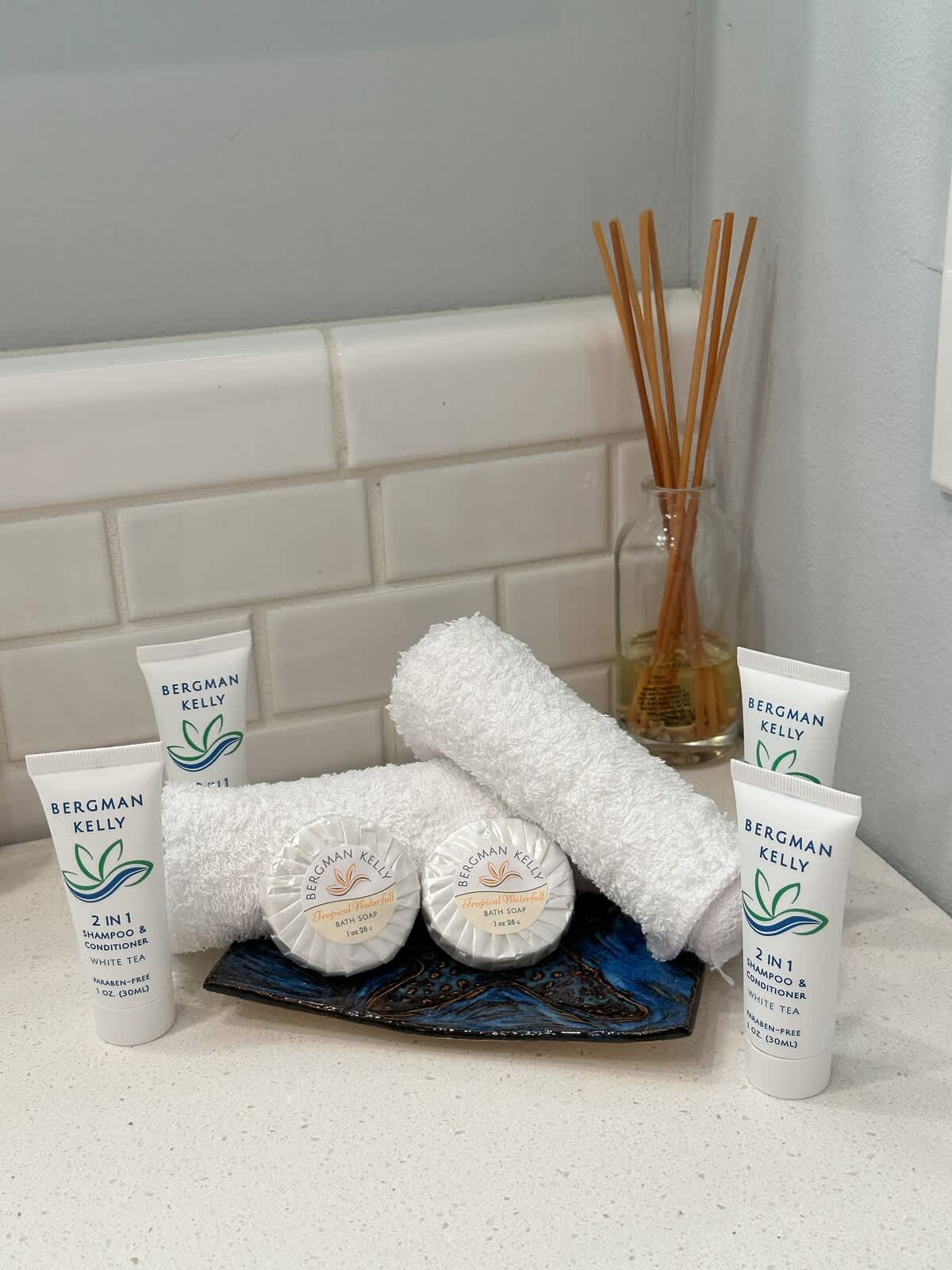 A close-up of bath amenities on a white countertop, including small white towels, two bars of soap, and two tubes of 2-in-1 shampoo and conditioner, arranged on a decorative blue and black tray.