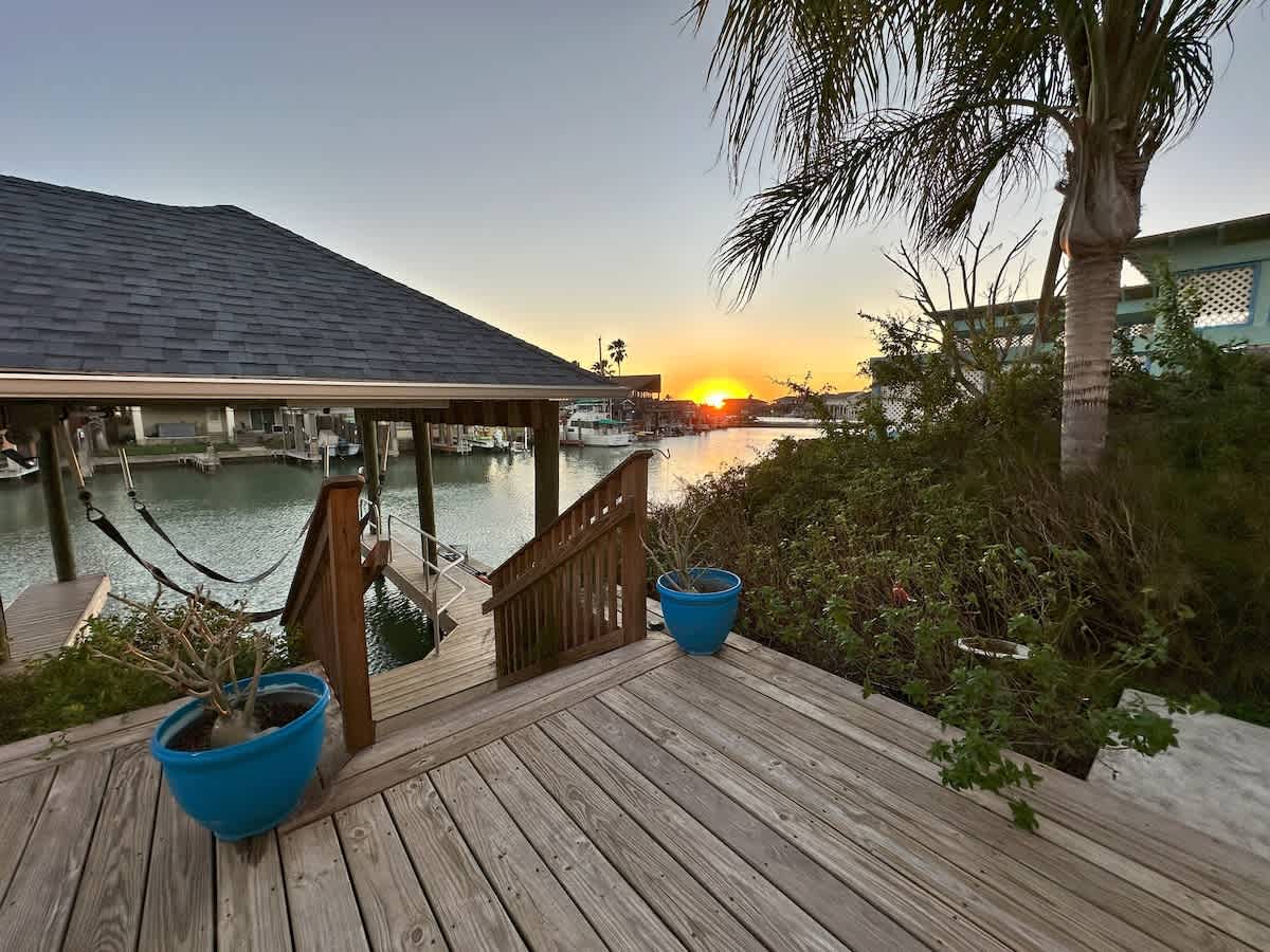 A spacious wooden deck with blue potted plants leads down to a boat dock on a tranquil canal, offering a picturesque view of the sunset over the water and distant palm trees.