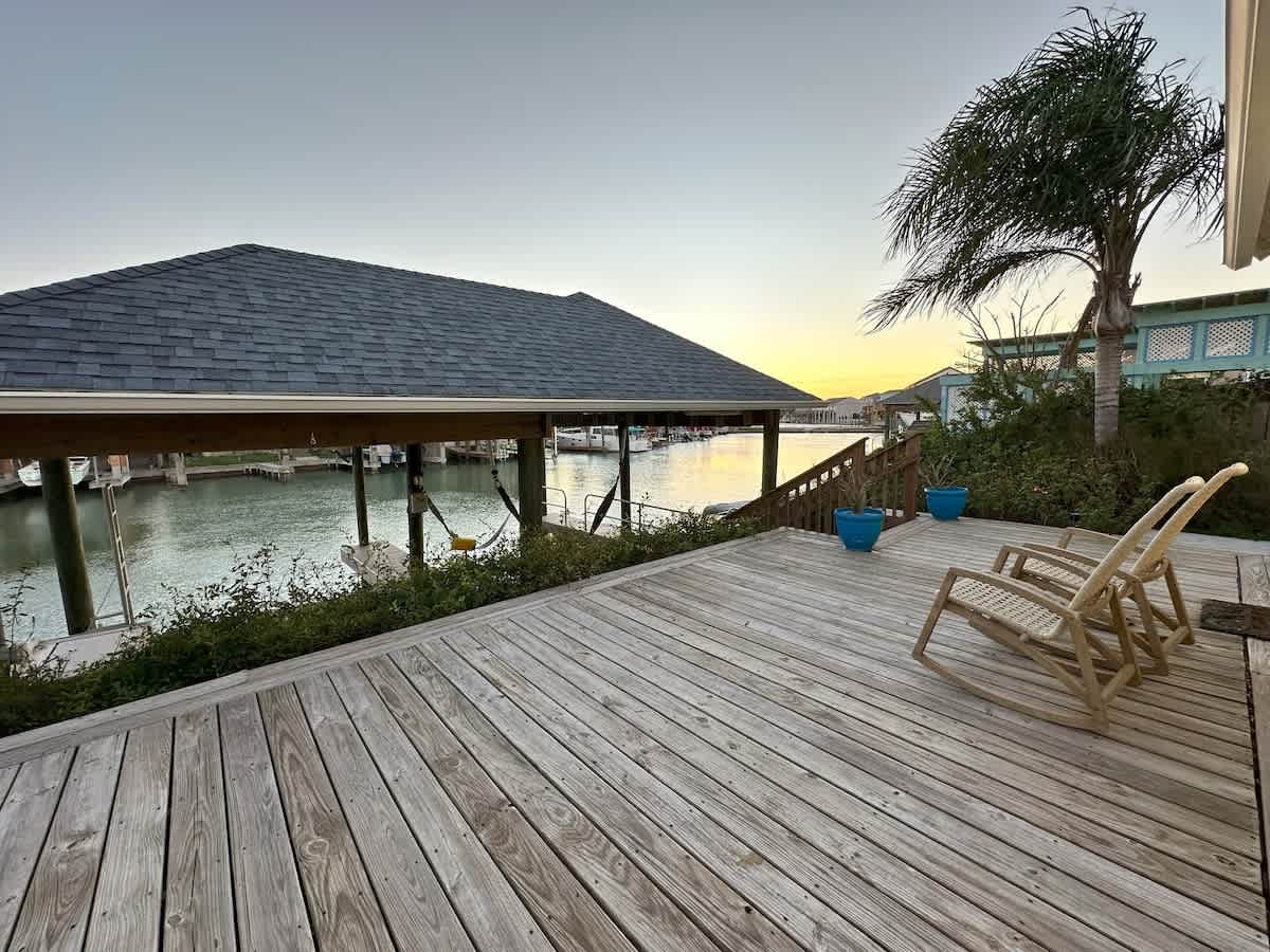 A wide wooden deck is furnished with two light-colored rocking chairs and blue potted plants, providing an expansive view of the covered boat dock and the scenic canal at sunset.