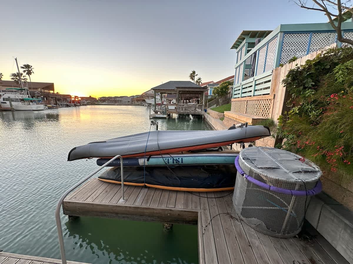 The end of a wooden boat dock is equipped with two kayaks or paddleboards and a round mesh container, set against the backdrop of the canal and a vibrant sunset horizon.