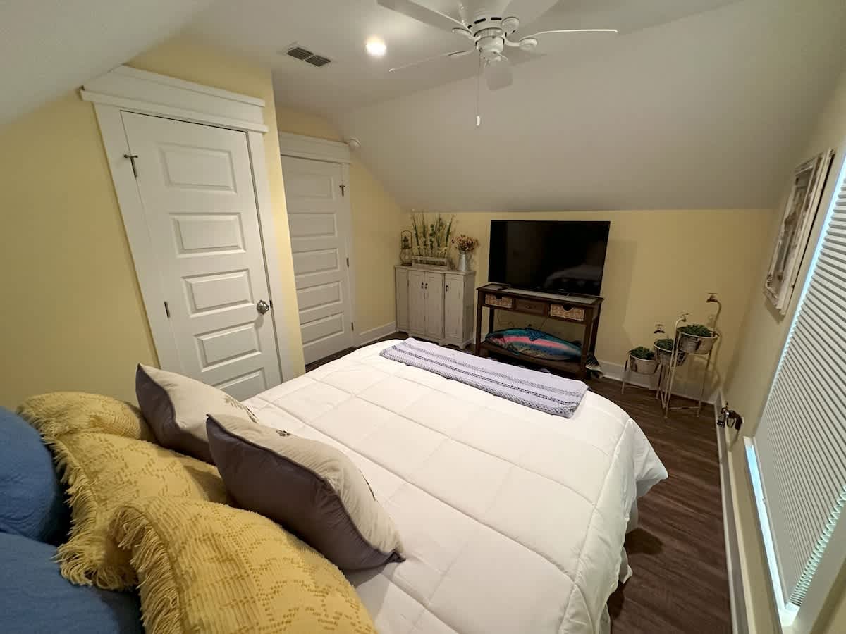 View of a cozy bedroom with light yellow walls and a sloped ceiling, showing a white bed with blue and yellow pillows, a wall-mounted TV on a console, and two white closet doors. Dark wood-look flooring is visible.