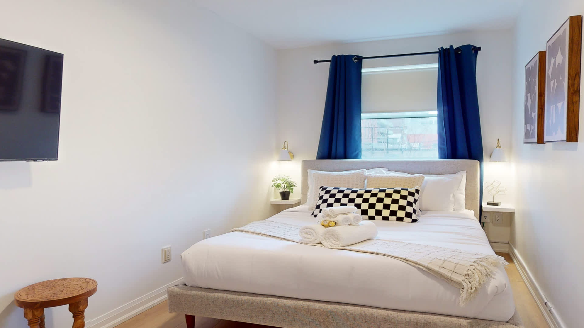 View of the second bedroom featuring a queen bed with white linens and a checkered accent pillow. A wall-mounted television is visible on the left, opposite the window with vibrant blue curtains, creating a comfortable retreat.