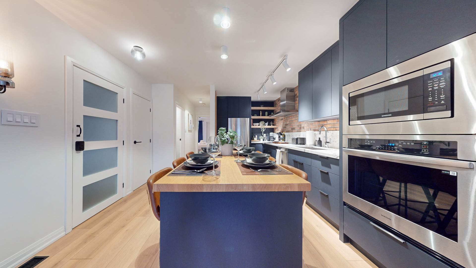 View from the kitchen island dining area towards a bright hallway with light wood floors and white doors featuring frosted glass panels. The dark blue kitchen cabinetry and brick wall are visible on the right, creating an open and modern feel.