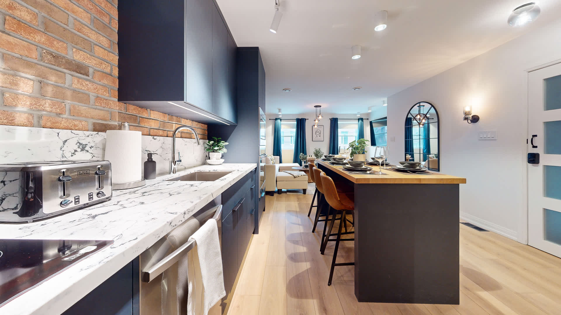 View of the kitchen sink area with white marble countertops, a stainless steel sink, and a toaster. Dark blue cabinetry lines the wall, and a brick accent wall is visible, connecting to the open-concept living space.
