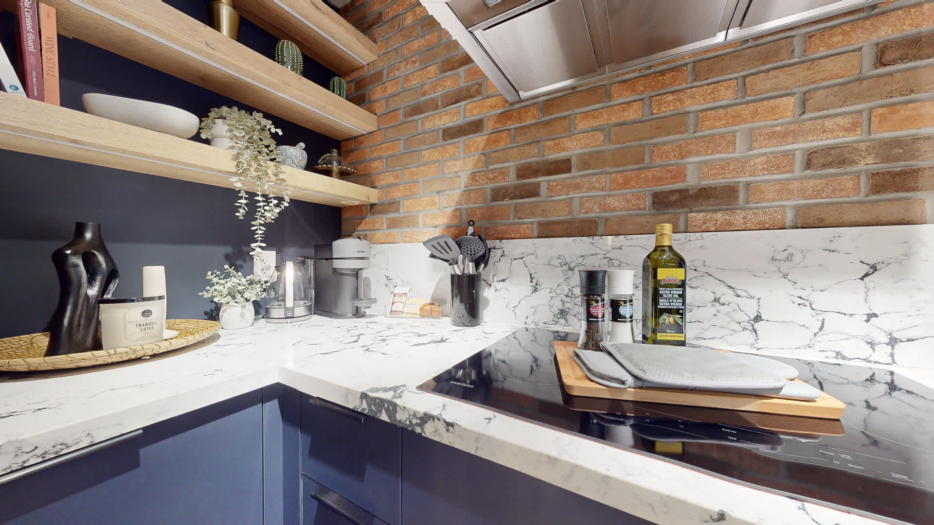 Corner of the kitchen showcasing open wooden shelves adorned with decorative items, above white marble-patterned quartz countertops. A brick accent wall and dark blue lower cabinets contribute to the modern industrial aesthetic.