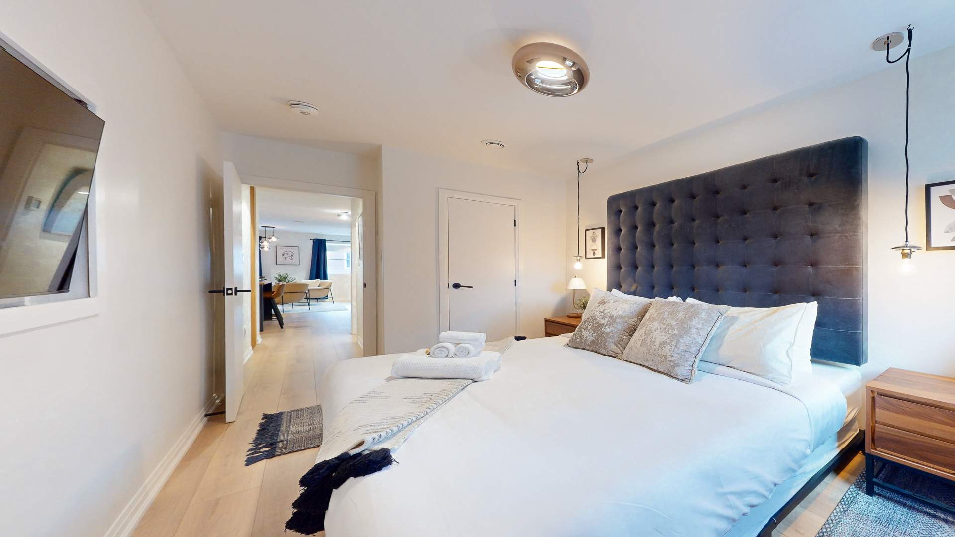 Elegant bedroom featuring a large grey tufted headboard, crisp white bedding, and light wood floors. A wall-mounted television and a glimpse of a hallway leading to other rooms are visible, creating a spacious feel.
