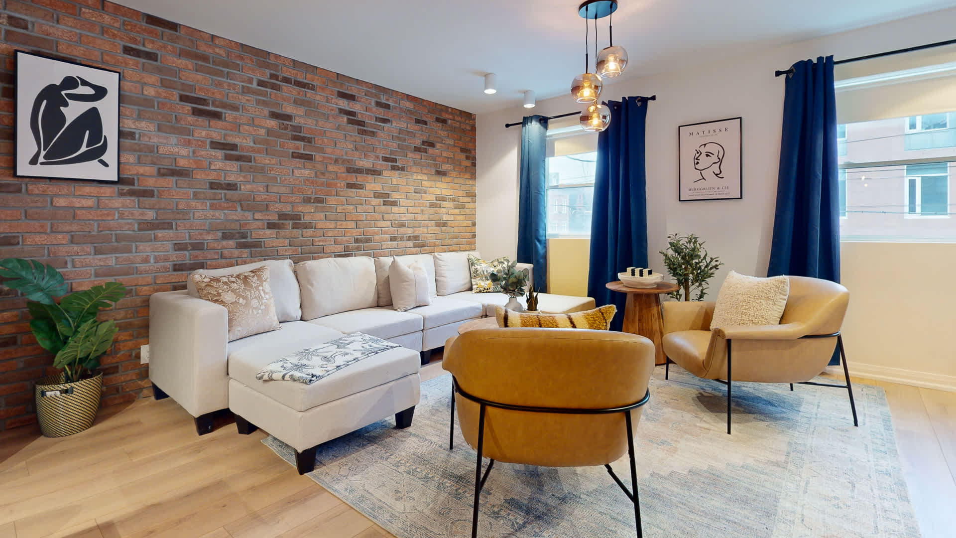 Cozy living room with a light-colored sectional sofa, two mustard yellow armchairs, and a round wooden coffee table. A brick accent wall provides a rustic touch, complemented by blue curtains and modern pendant lighting.