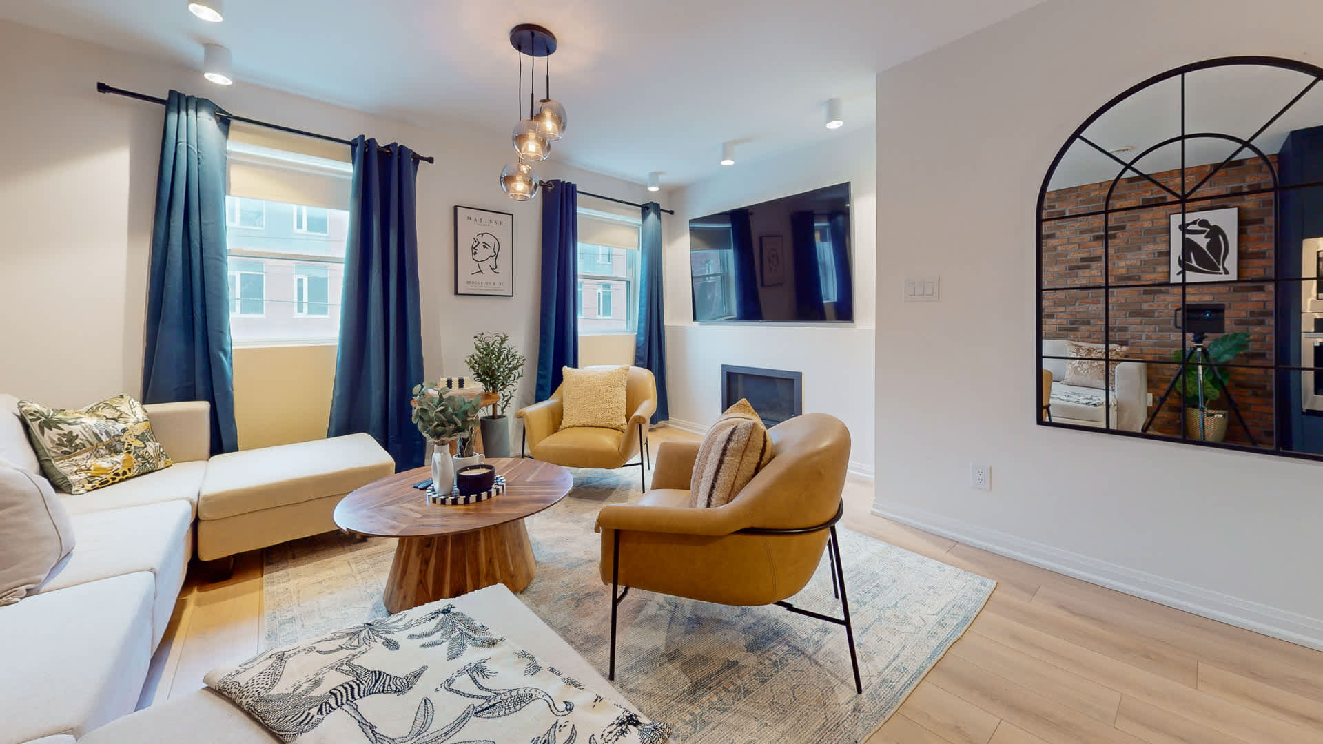 Bright living room with a wall-mounted TV above a modern fireplace, flanked by blue-curtained windows. A large arched mirror reflects the brick accent wall, adding depth to the space with a sectional and armchairs.