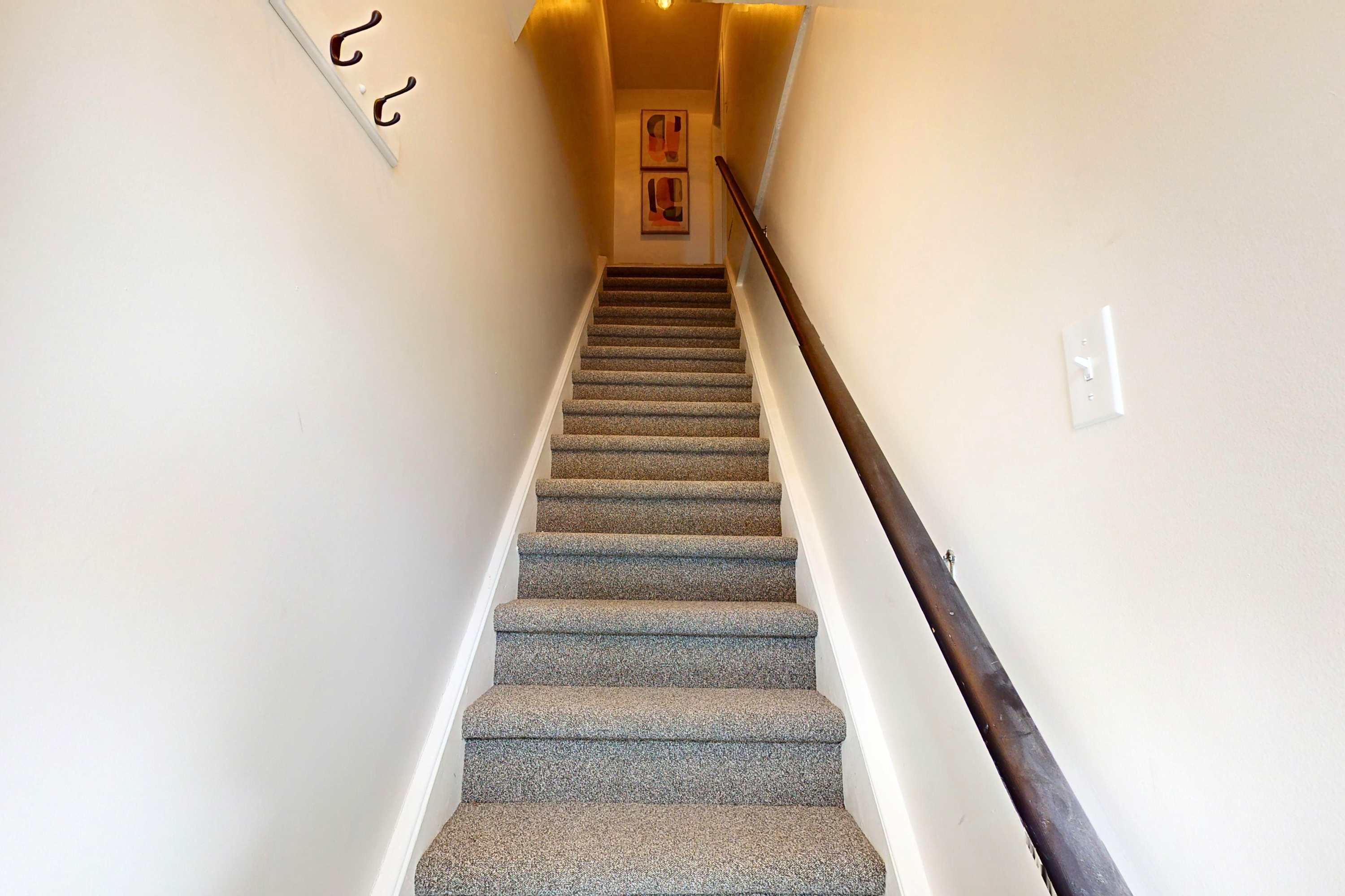 A carpeted staircase with a dark wooden handrail ascends towards a landing with two framed artworks. The walls are painted a light neutral color.