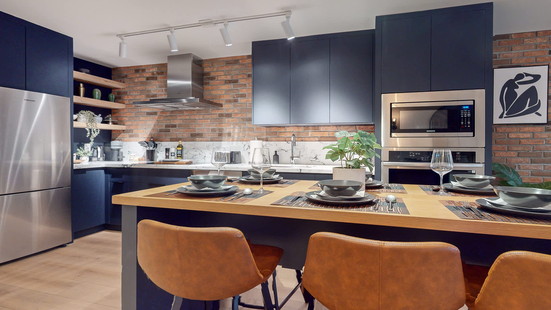 Contemporary kitchen with dark blue flat-panel cabinets, a light wood island with seating for four, and a prominent brick accent wall. Integrated stainless steel appliances, including a built-in oven and microwave, are set against white marble-patterned countertops.