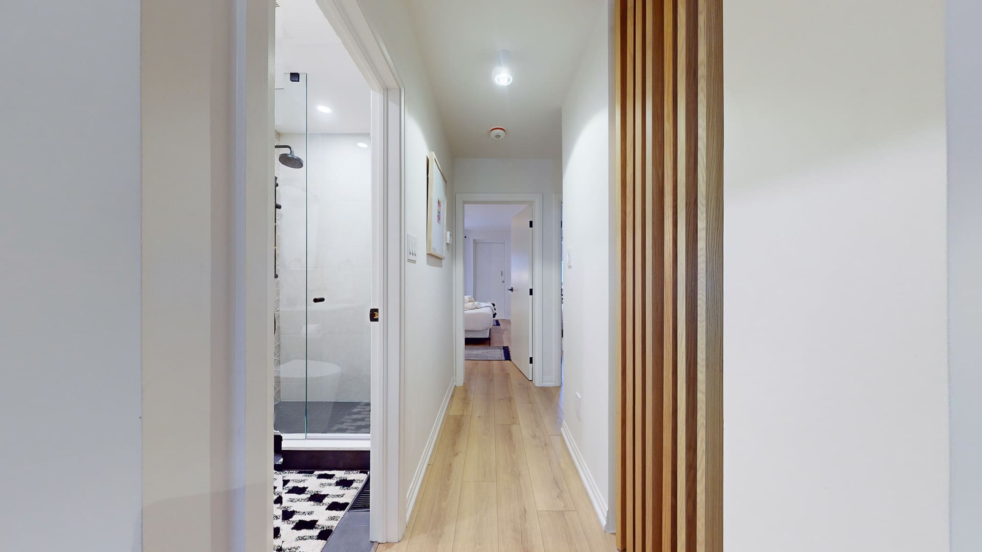 Bright hallway with light wood flooring, leading past a modern bathroom with a glass shower enclosure on the left. A decorative wooden slat wall panel adds texture to the right, enhancing the contemporary design.