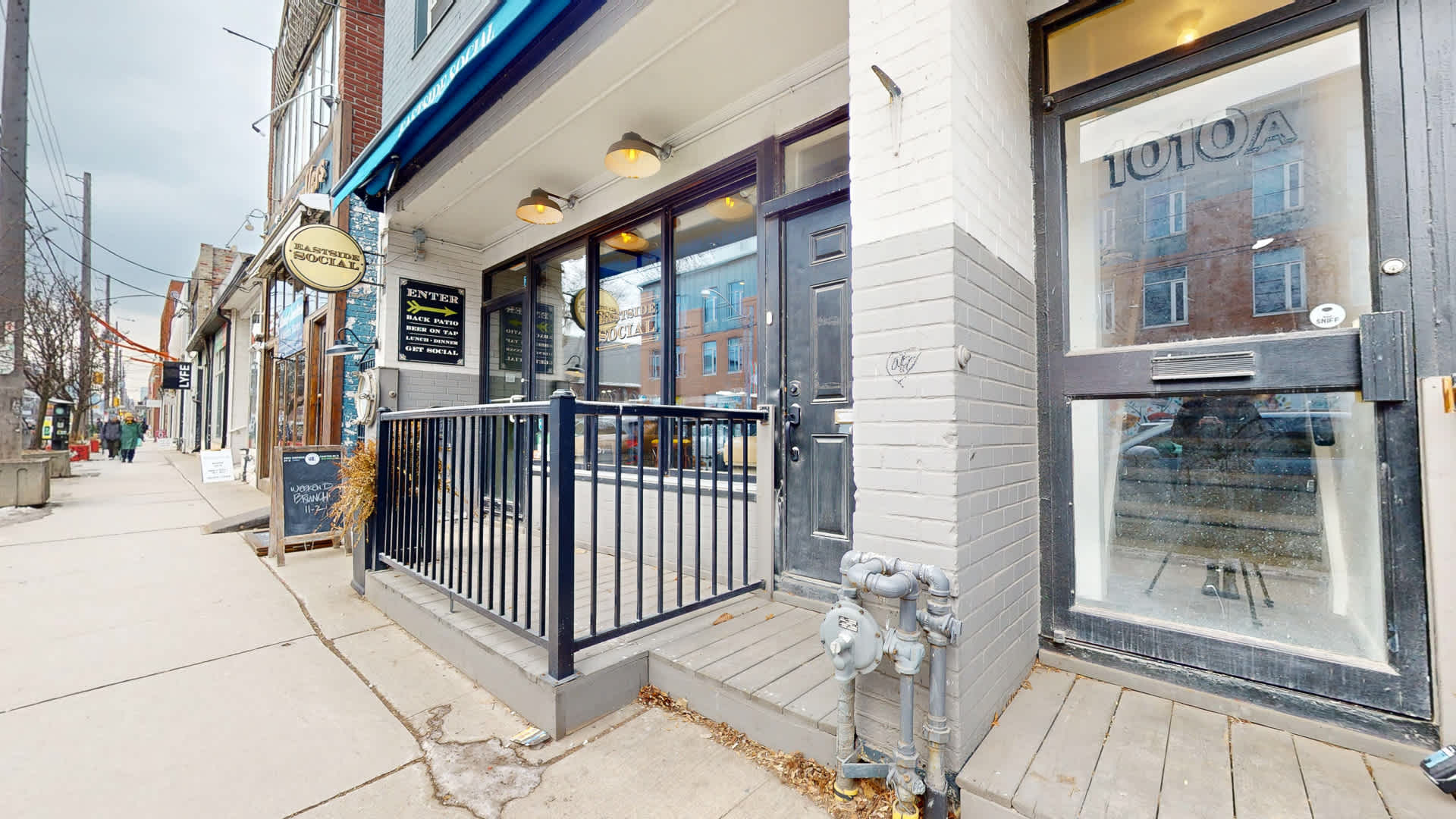 Wider street-level view of the commercial storefront, showing a brick building with large windows and a black door. A small wooden deck with a black railing leads to the entrance, with a street and other buildings visible in the background.