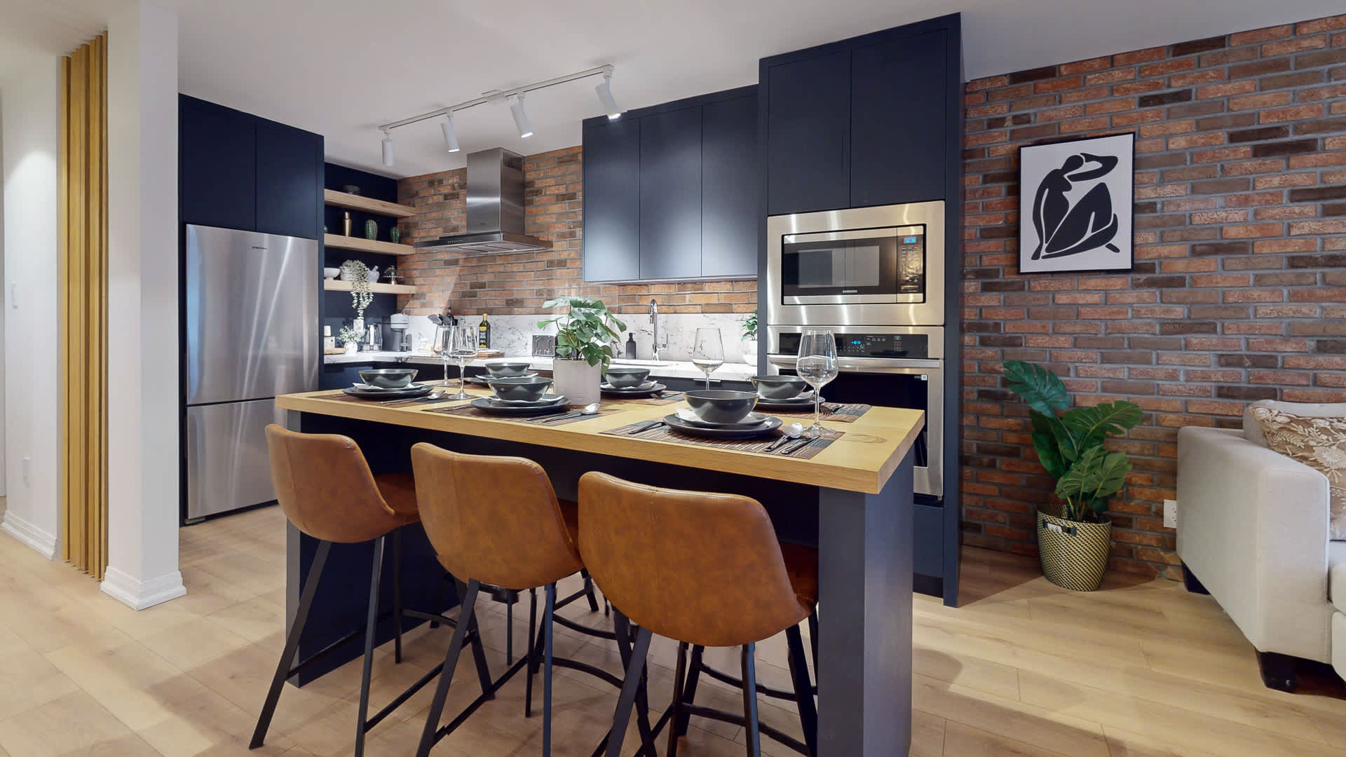 Modern kitchen featuring dark blue flat-panel cabinetry, a light wood island serving as a dining table with bar stools, and a striking brick accent wall. Stainless steel appliances and white marble-patterned quartz countertops complete the contemporary design.