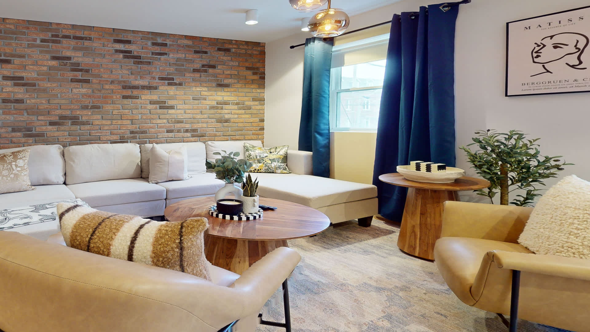 Inviting living room showcasing a light sectional sofa, two mustard armchairs, and a round wooden coffee table with decorative items. A rustic brick wall and blue drapes add character, with natural light filtering in.
