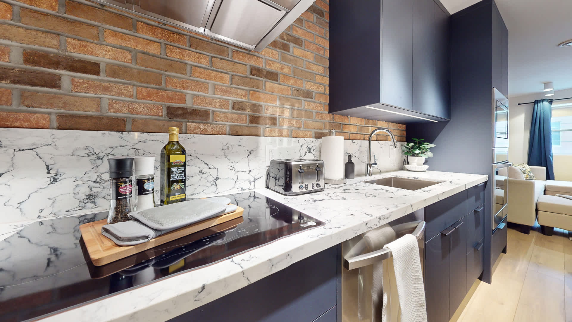 Detailed view of a modern kitchen countertop featuring white marble-patterned quartz and a sleek black induction cooktop. A stainless steel range hood, brick backsplash, and various kitchen essentials like a toaster and olive oil are visible.