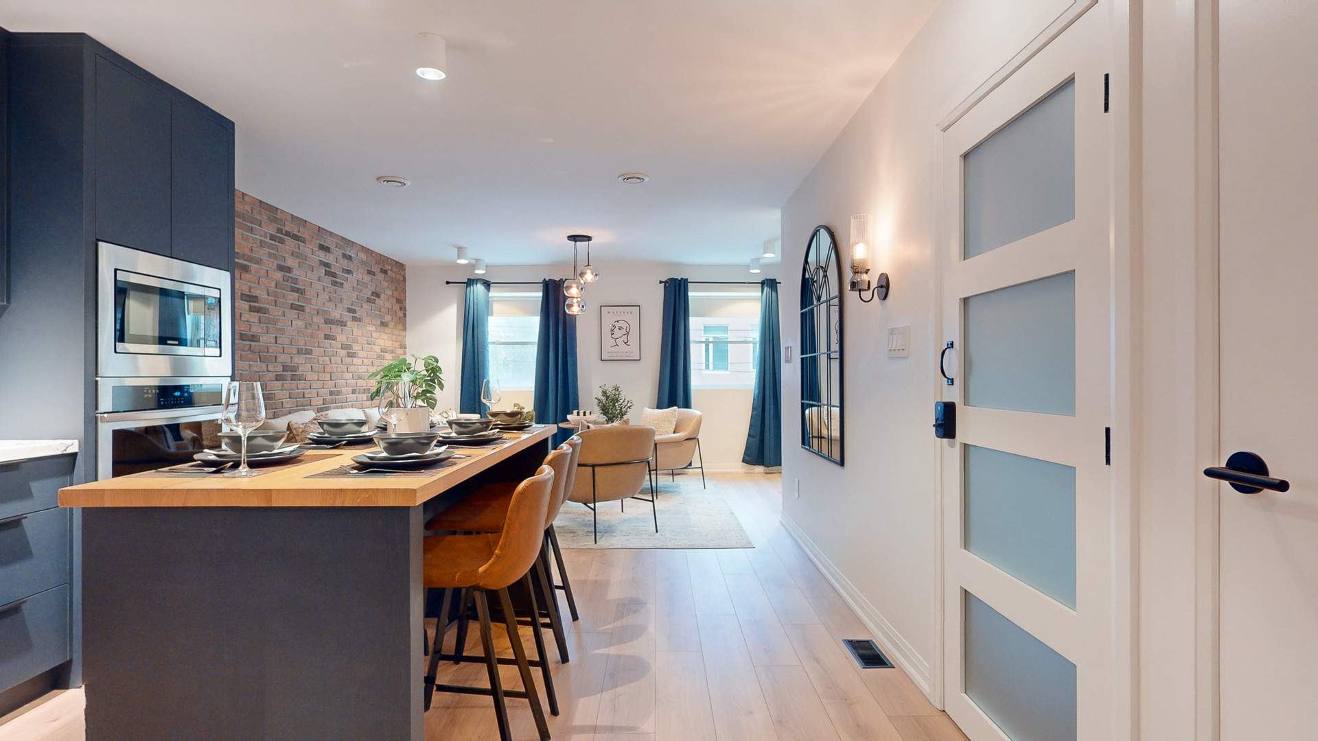 Open-concept living space with a kitchen island in the foreground, leading to a cozy living room featuring light-colored armchairs, a patterned rug, and blue curtains. Hardwood floors seamlessly connect the kitchen and living areas.