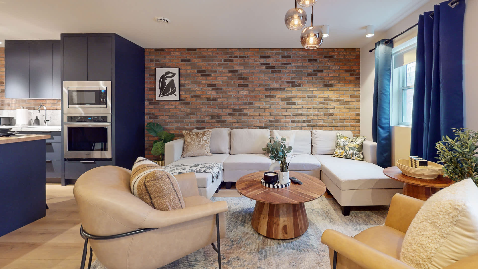 Open-concept living room and kitchen area, featuring a light sectional sofa and armchairs facing a brick accent wall. Dark blue kitchen cabinetry and built-in appliances are visible in the background, connecting the spaces.