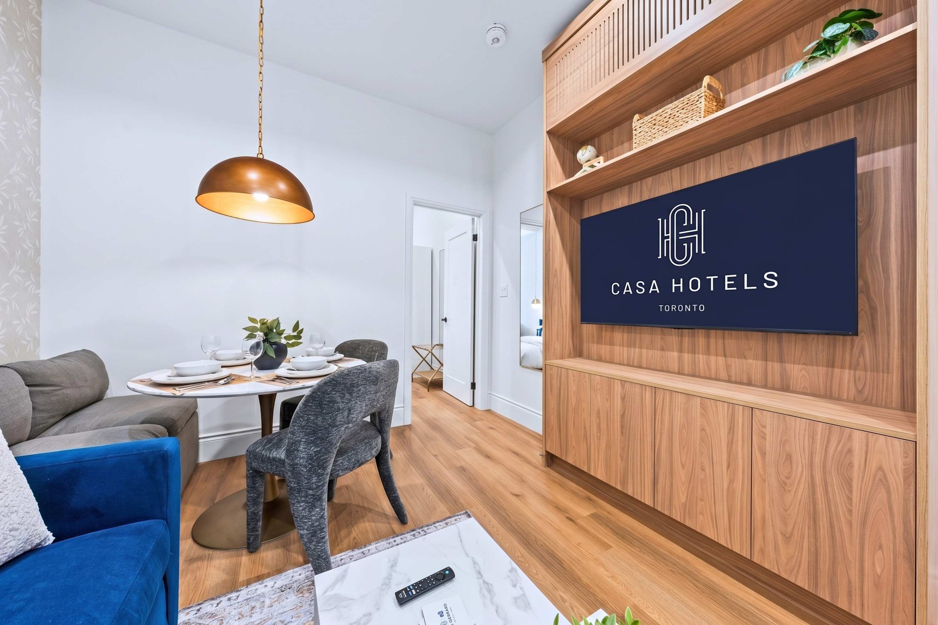 Living area featuring a blue sofa, a round dining table with banquette seating, and a custom wooden media unit with open shelving. A large flat-screen TV displaying the 'CASA HOTELS TORONTO' logo is integrated into the wooden unit.