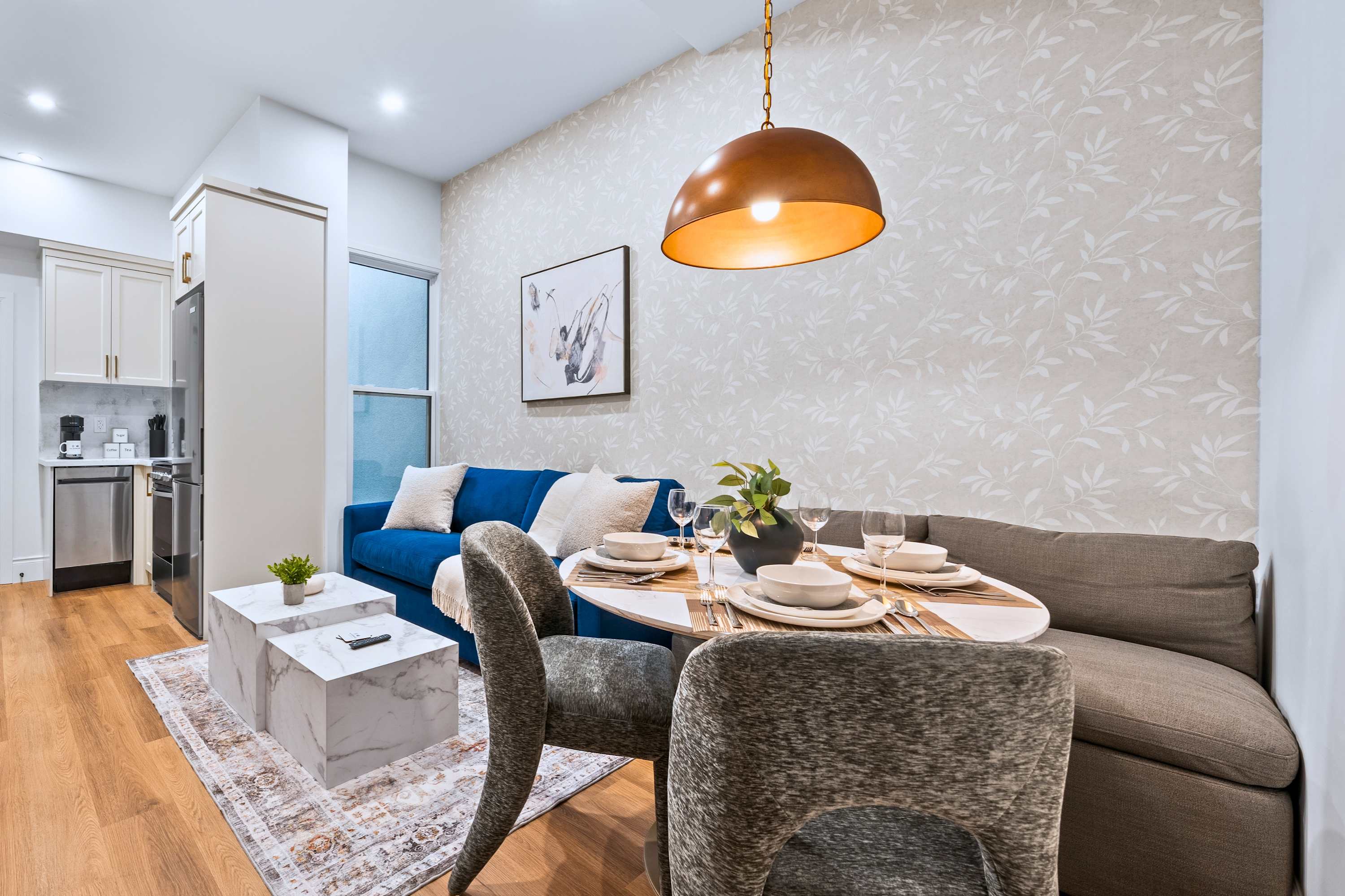 Cozy living and dining area with a blue sofa, a white marble coffee table, and a round dining table with banquette seating and two upholstered chairs. A modern abstract artwork hangs on the wallpapered accent wall, and a kitchen is visible in the background.