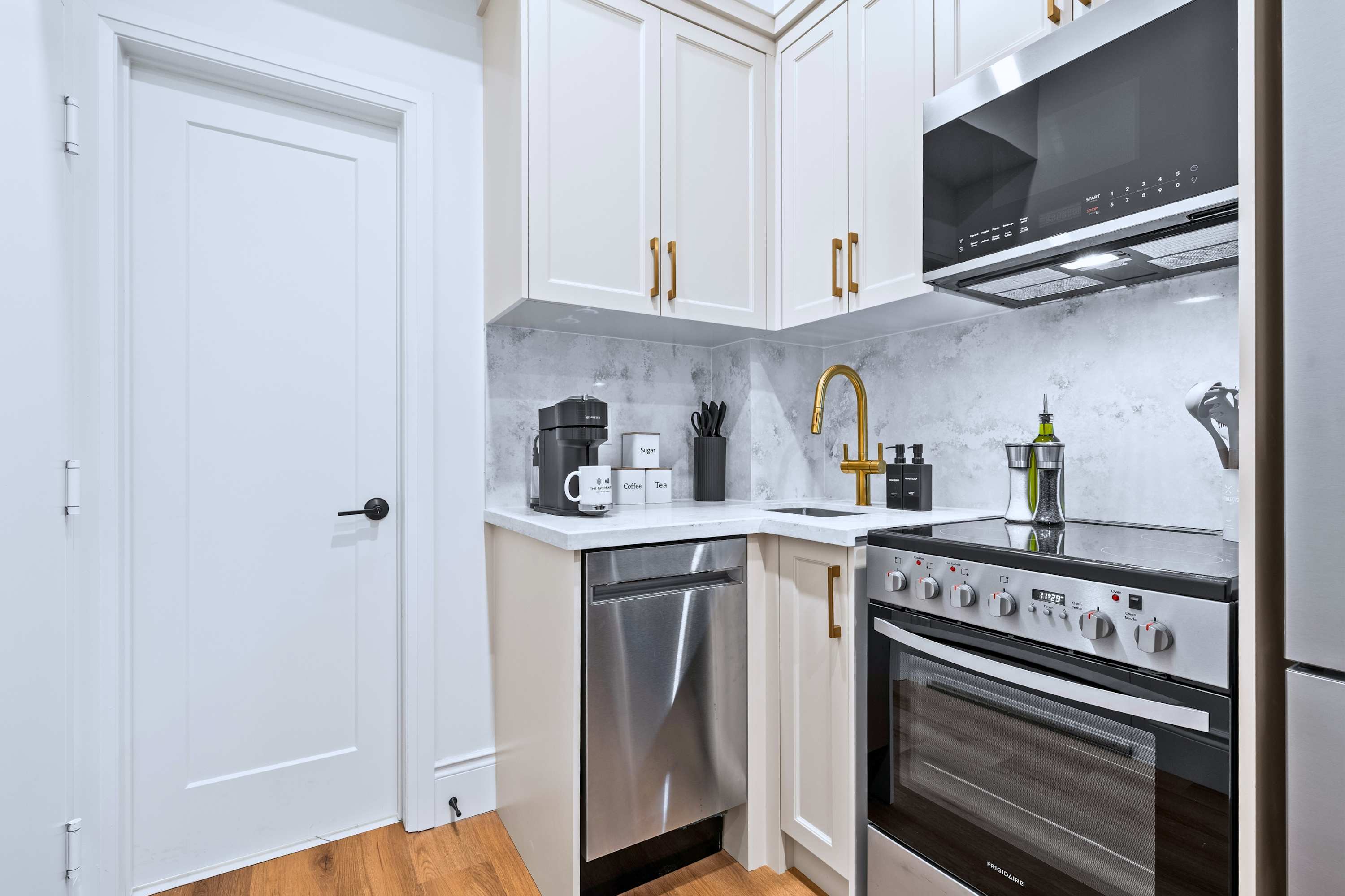 Compact and modern kitchen with light cream shaker cabinets, gold hardware, white quartz countertops, and a grey marble-patterned backsplash. It features stainless steel appliances including a dishwasher, oven/stove, and microwave, along with a gold faucet and coffee maker.