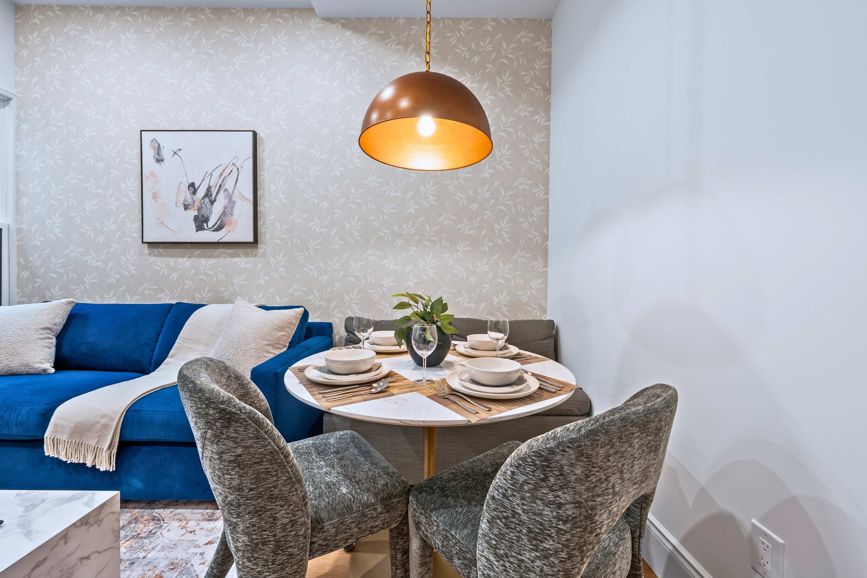 Cozy living and dining area featuring a blue velvet sofa, a round white marble-top dining table with two upholstered chairs, and a built-in banquette, all under a copper dome pendant light. The wall is adorned with light-patterned wallpaper and a framed abstract art piece.