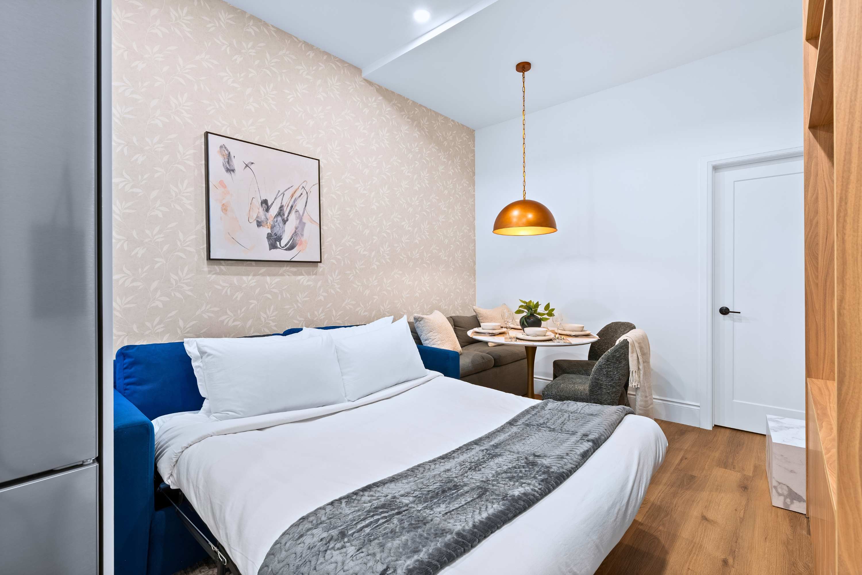 The living area transforms into a bedroom with a blue sofa bed pulled out, revealing a white bed with a grey throw. The space includes a round dining table with chairs and a banquette, illuminated by a copper pendant light, and features patterned wallpaper on one wall.