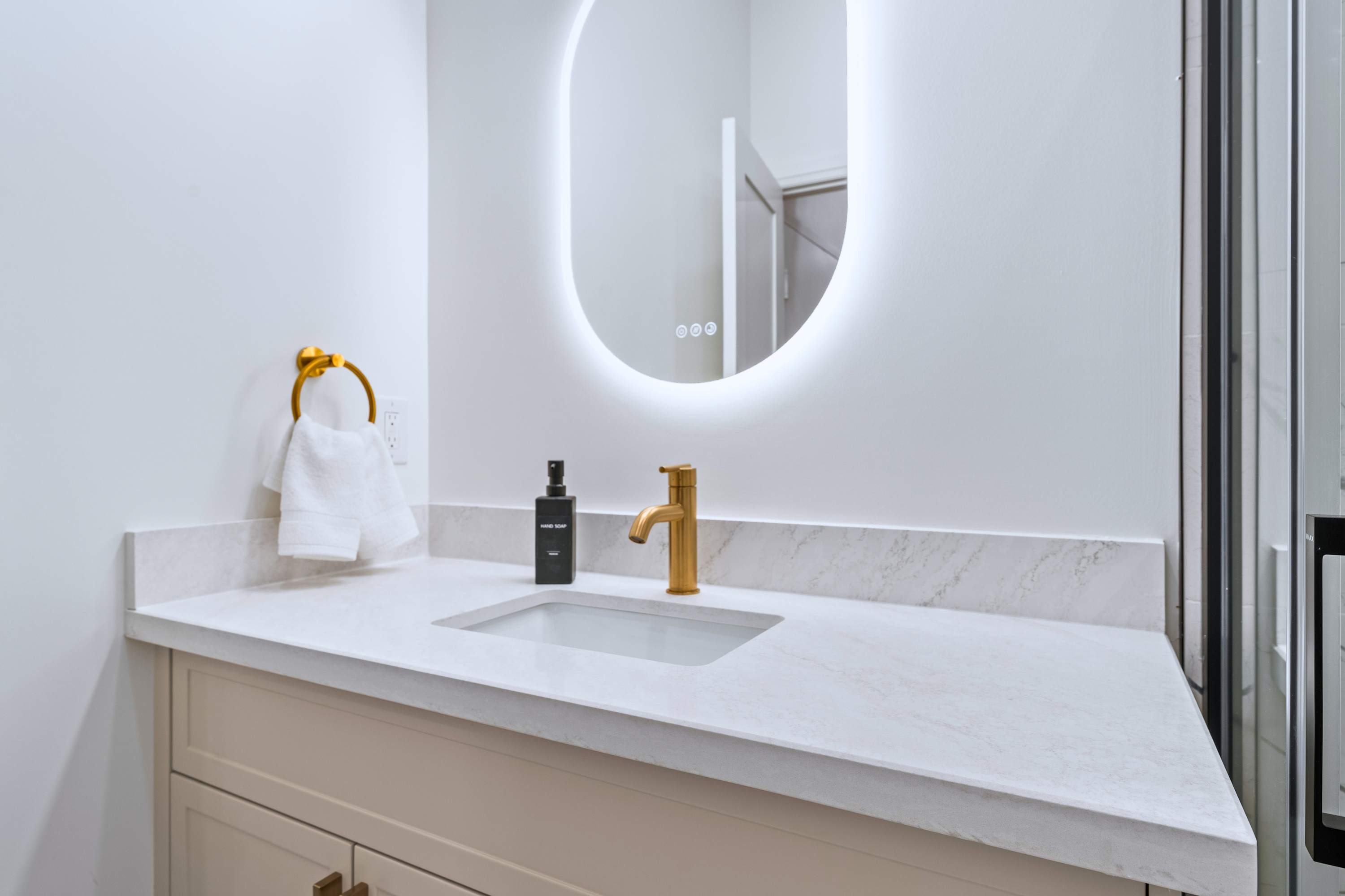 Close-up of the bathroom vanity, featuring a white countertop with an integrated sink, a gold faucet, and a gold towel ring with a white hand towel. The large oval illuminated mirror reflects the opposite wall.