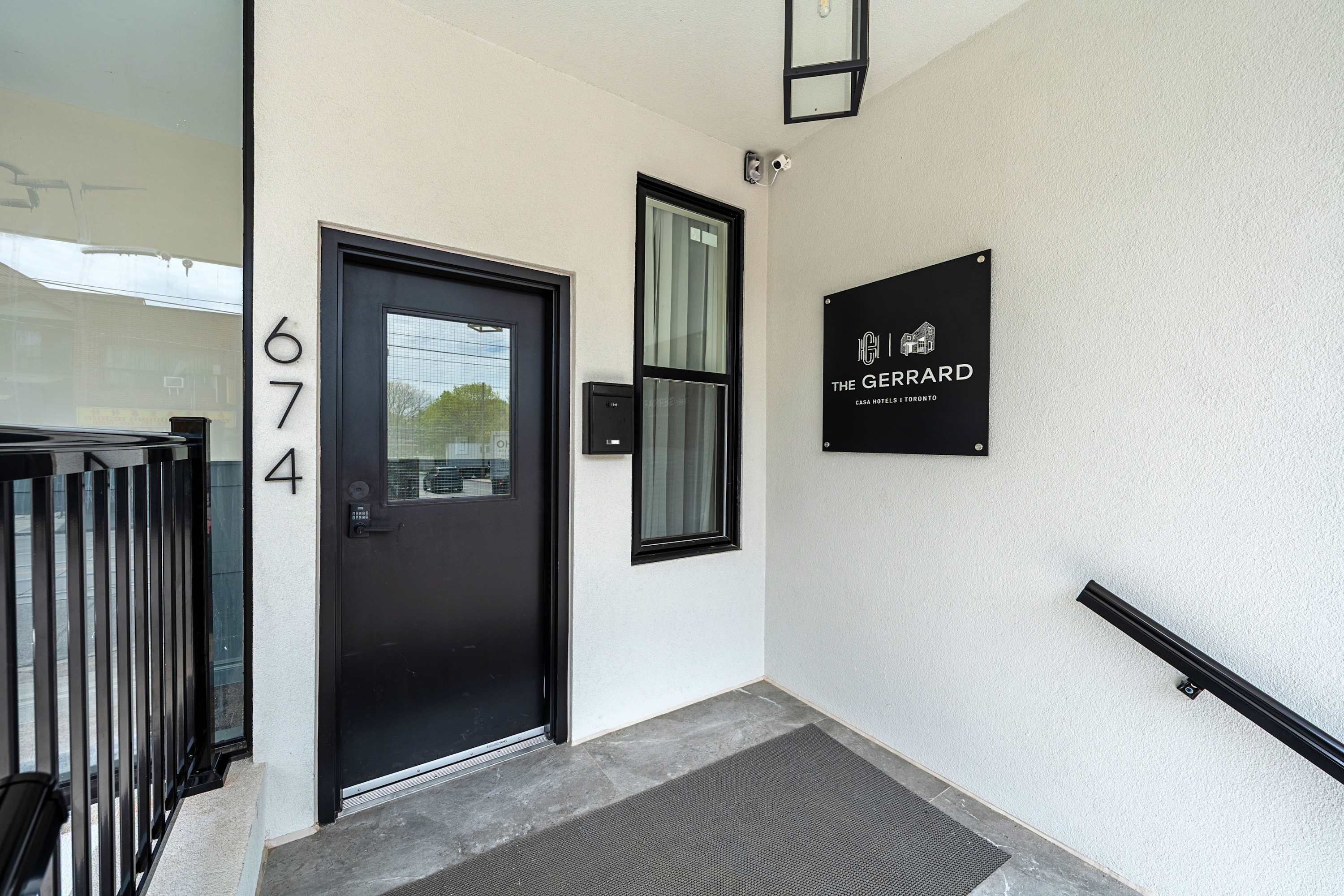 A close-up of the hotel entrance shows a black door with a modern lock, a narrow window, and a black sign for "THE GERRARD CASA HOTELS | TORONTO" mounted on the white wall.
