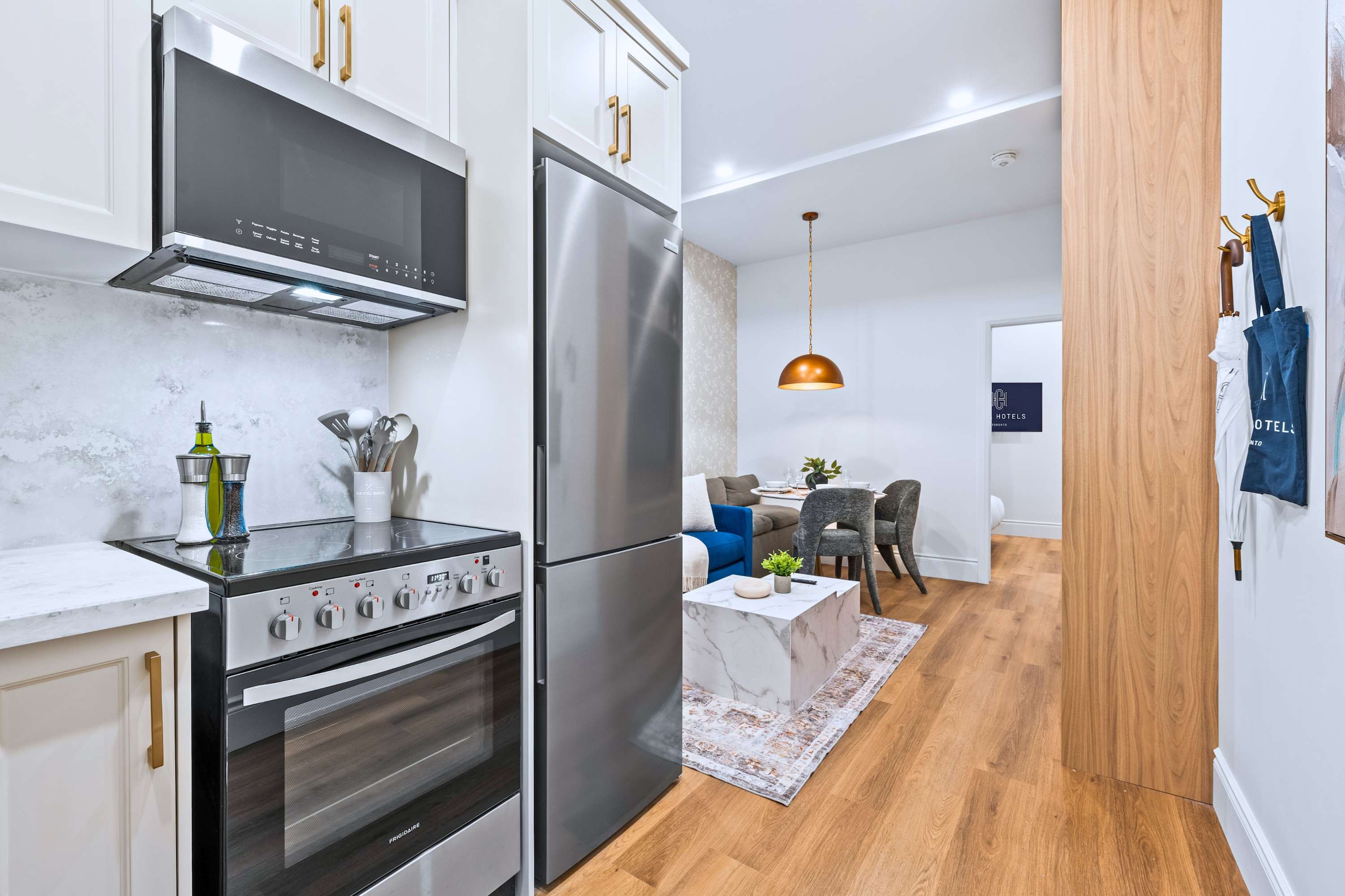 View of the compact kitchen from the hallway, showcasing stainless steel appliances including a refrigerator, oven, and microwave, alongside light cream cabinets and a marble-patterned backsplash. The hallway leads to the living/dining area, visible in the background.