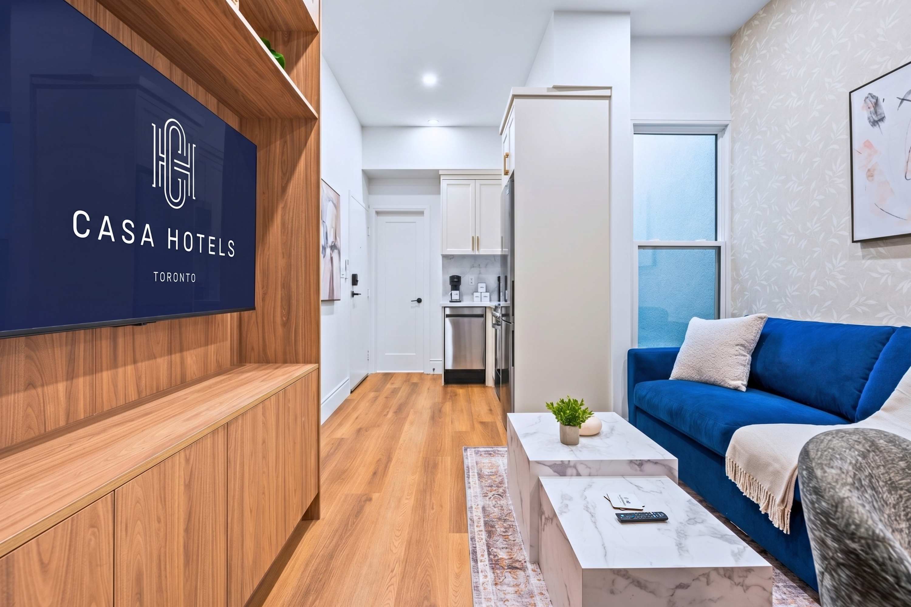 Modern living room with a vibrant blue velvet sofa, two nested marble-top coffee tables, and a large wall-mounted TV displaying 'CASA HOTELS TORONTO'. The room features light hardwood floors, a patterned rug, and a built-in wooden shelving unit.