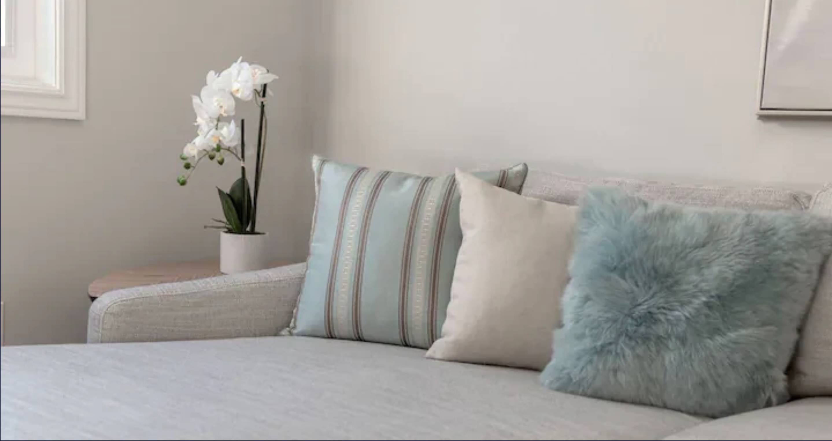 Close-up view of a light grey sectional sofa adorned with striped, plain, and fluffy blue throw pillows. A white orchid plant on a side table adds a touch of elegance next to a window.