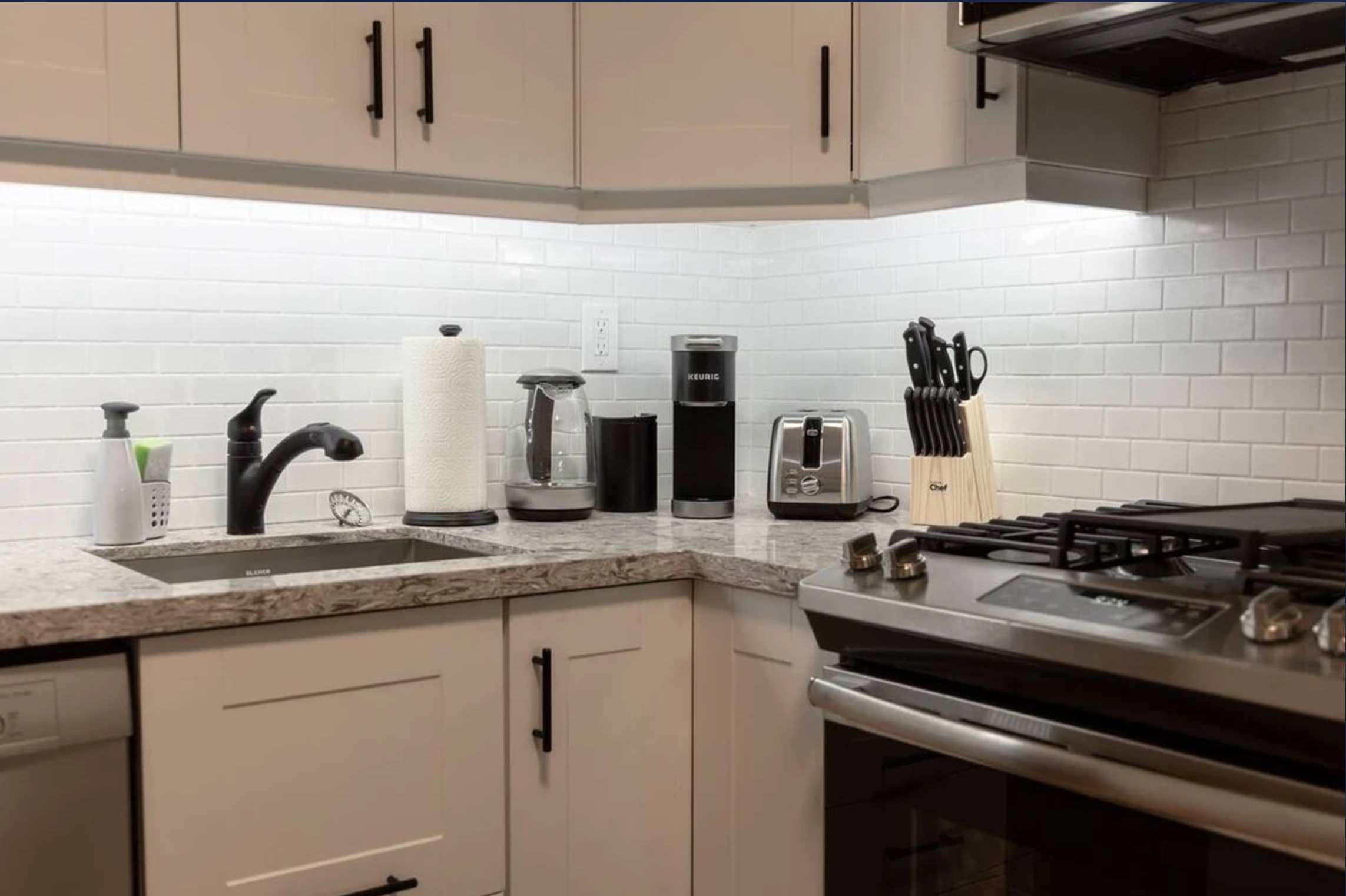 Modern kitchen featuring white subway tile backsplash, light gray granite countertops, and white shaker cabinets. Stainless steel appliances include a gas range, sink, toaster, and coffee maker, all illuminated by under-cabinet lighting.
