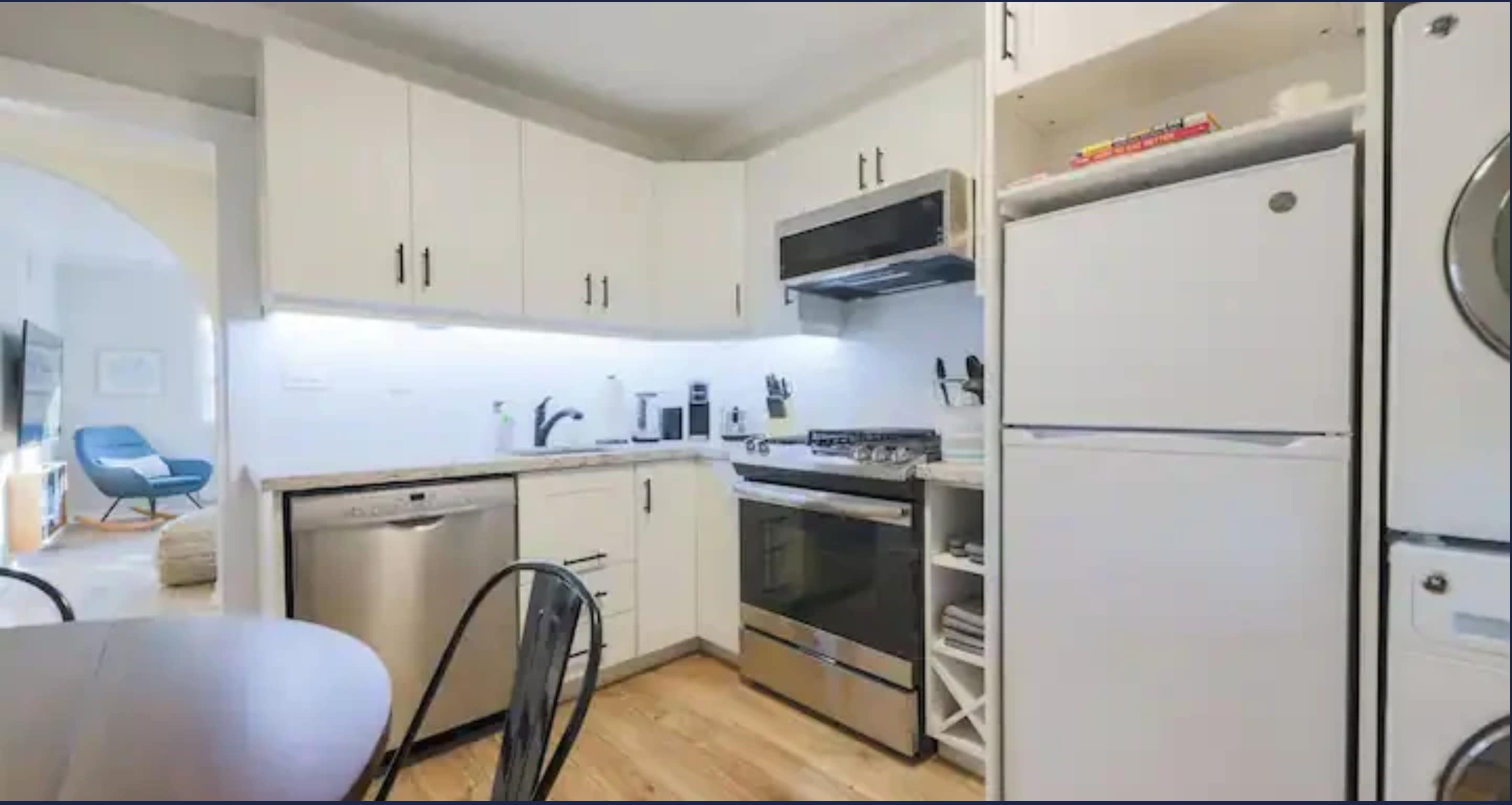 Wide shot of the modern kitchen with white shaker cabinets, stainless steel appliances including a dishwasher, oven, and microwave. A white refrigerator and stacked washer/dryer are visible on the right, with a dining table in the foreground.