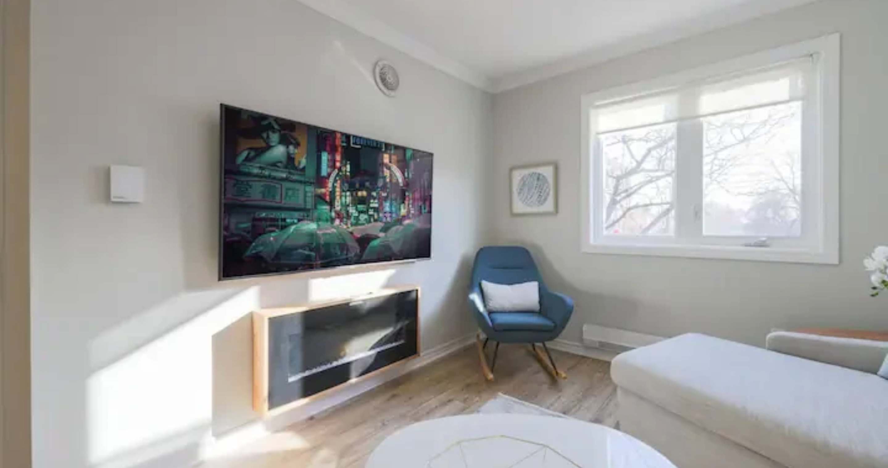 Living room with a large wall-mounted TV displaying a vibrant city scene, positioned above a modern electric fireplace. A blue rocking chair sits next to a window, and light hardwood floors extend throughout.