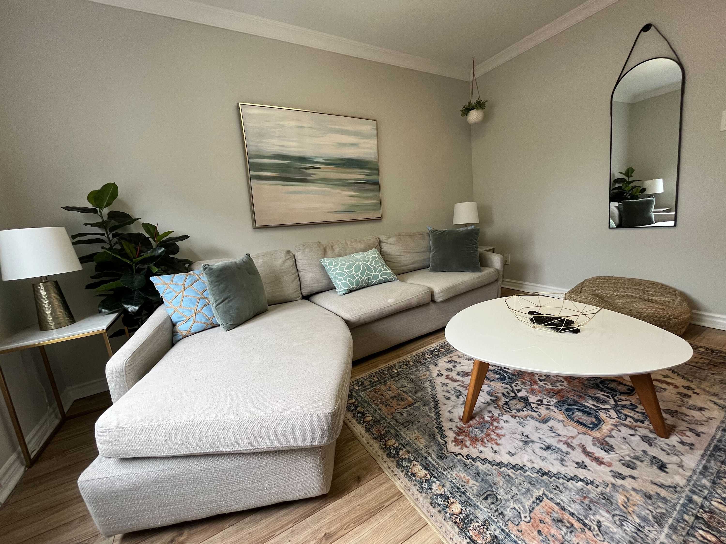 Stylish living room featuring an L-shaped sectional sofa with various throw pillows, a white oval coffee table, and a patterned area rug. A large abstract painting hangs above the sofa, and a full-length mirror is on the adjacent wall.